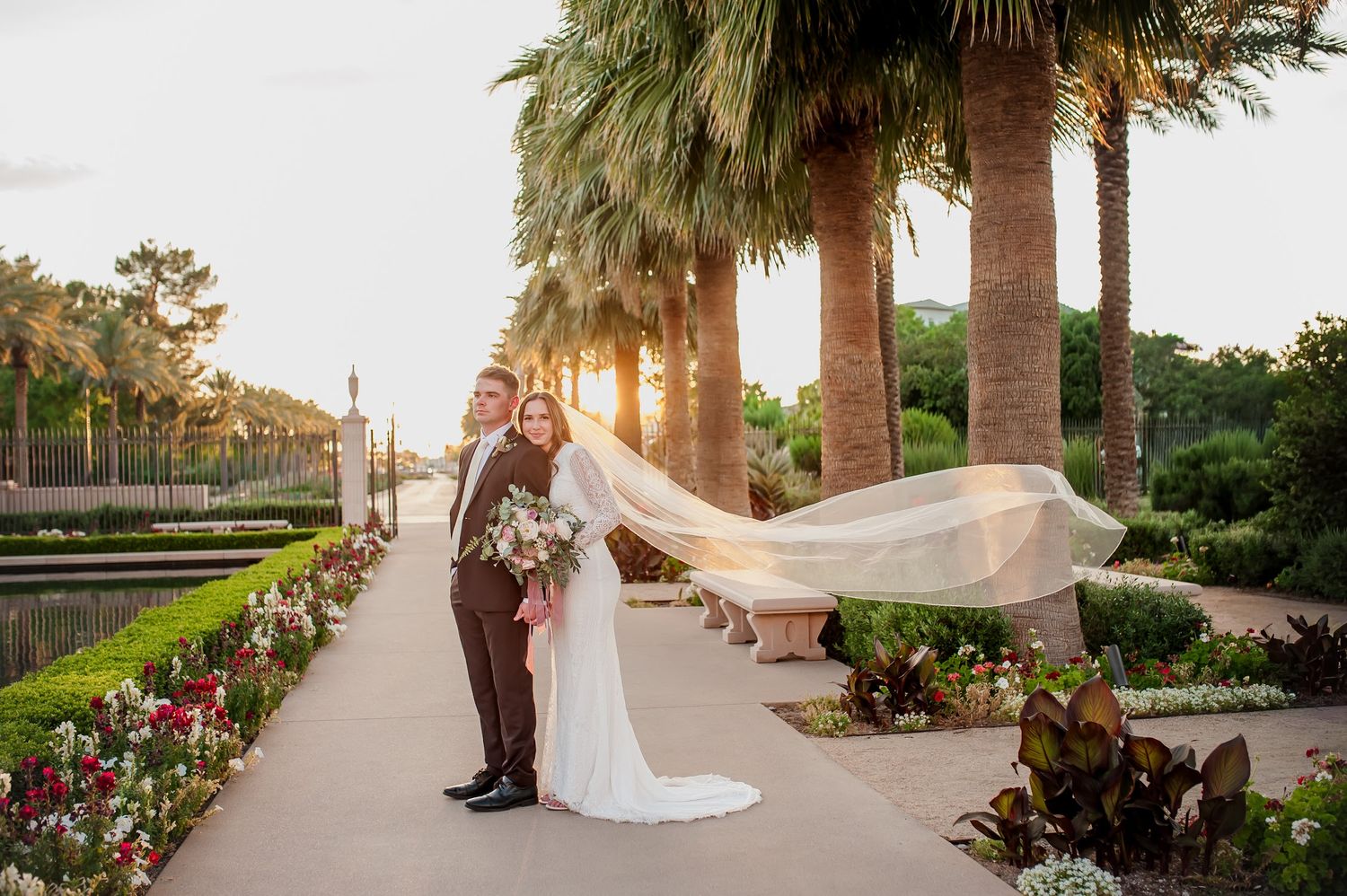 Bride and groom pose in a sunlit garden with palm trees, flowing veil caught in the breeze at golden hour.