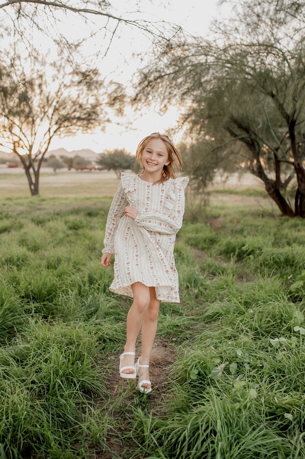 A young girl in a white floral dress walks through a lush green field at golden hour.