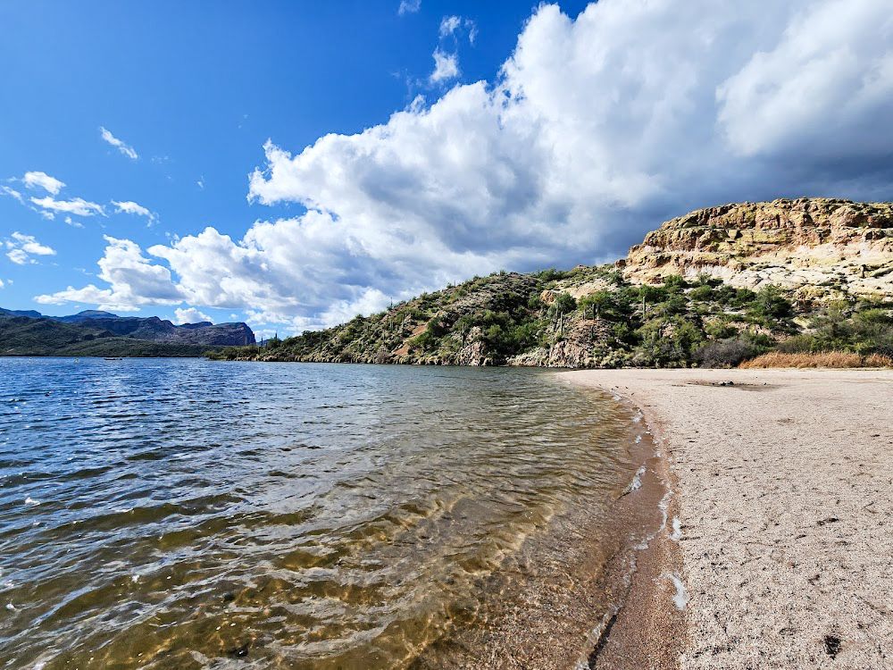 Serene desert lake with clear shallow waters, sandy beach, rocky cliffs, and dramatic cloudy blue sky in Arizona.