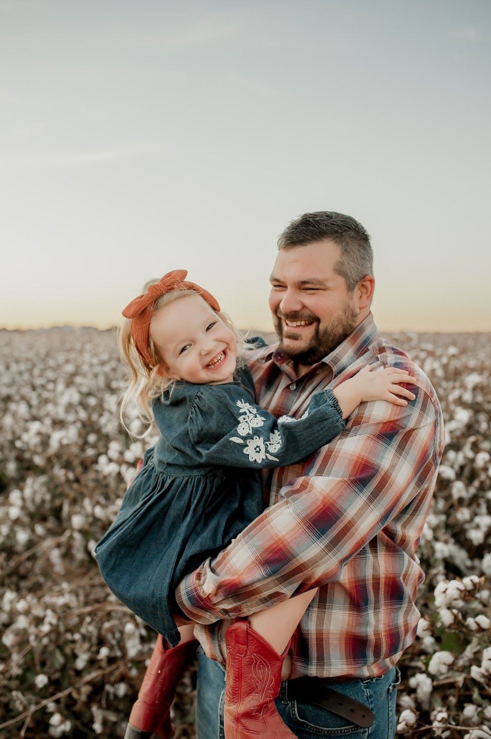 A smiling man holds a laughing little girl in a cotton field at sunset, both wearing plaid and denim outfits.