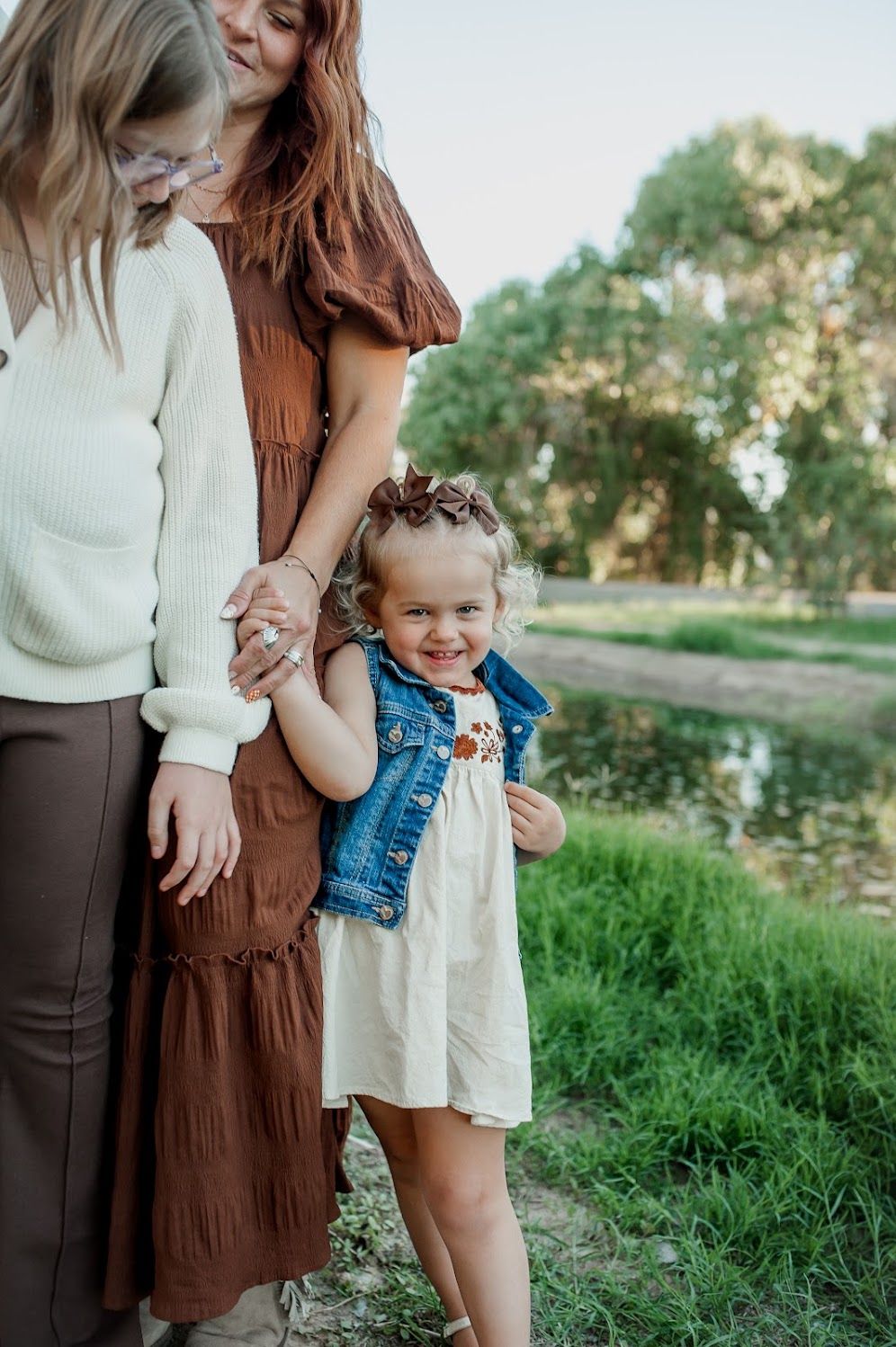 Two women and a smiling toddler girl in a denim vest pose outdoors near a pond surrounded by green trees.