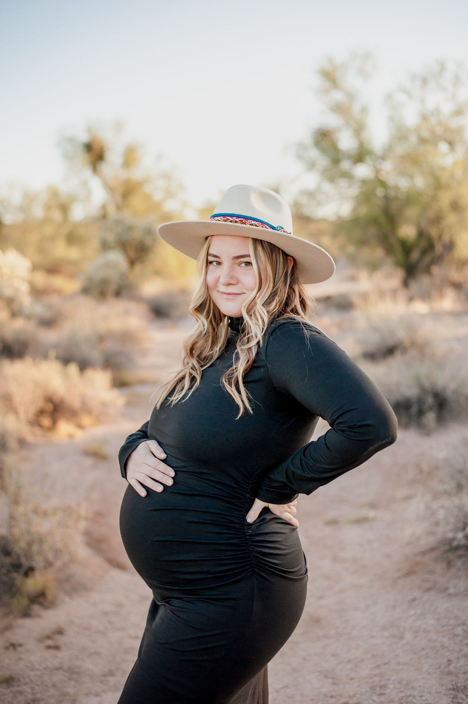 Expectant mother in black dress and beige hat poses in desert landscape during golden hour maternity photoshoot.