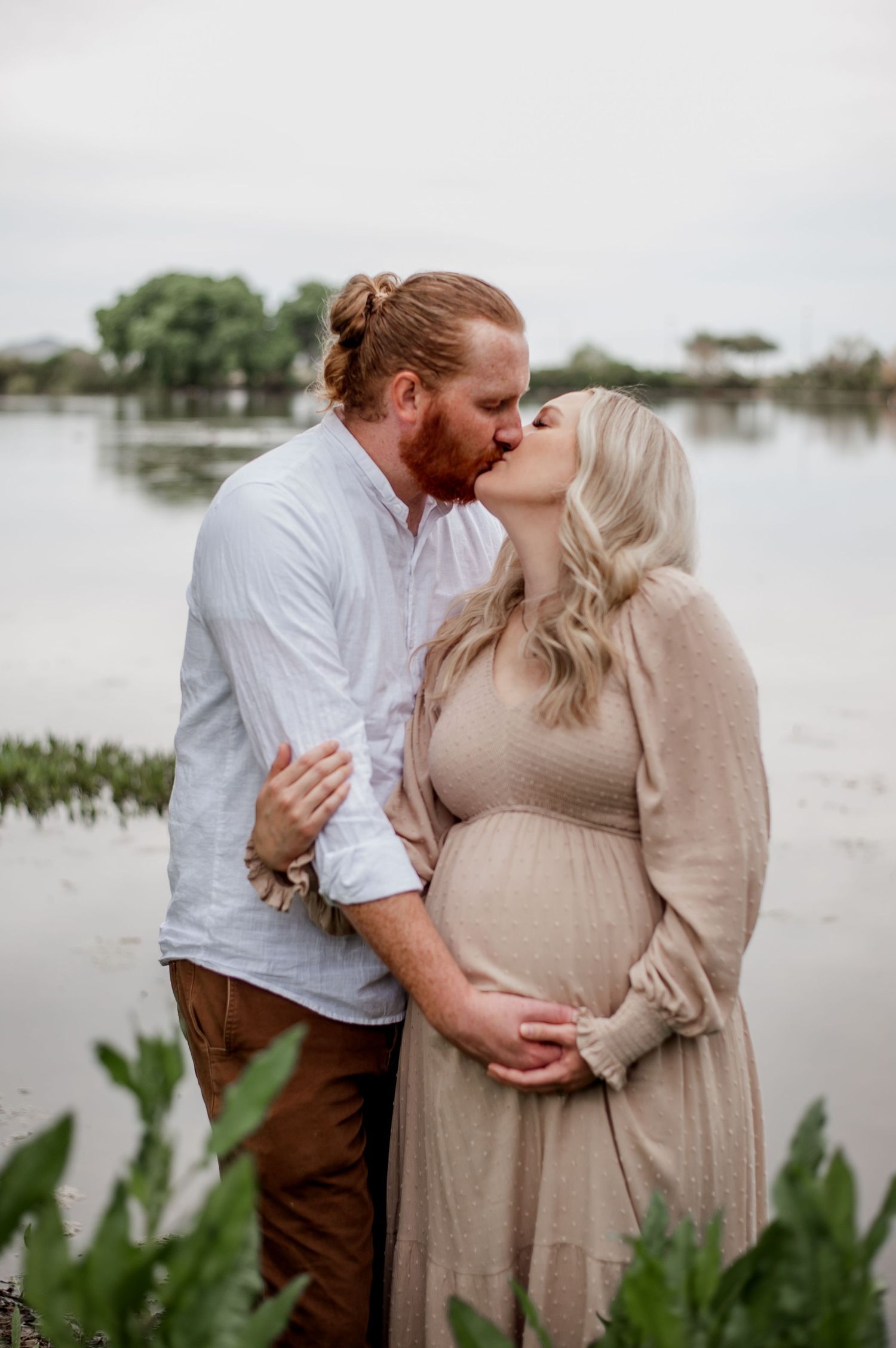 Maternity photoshoot by a lake featuring a couple in white and beige clothing sharing romantic moments.