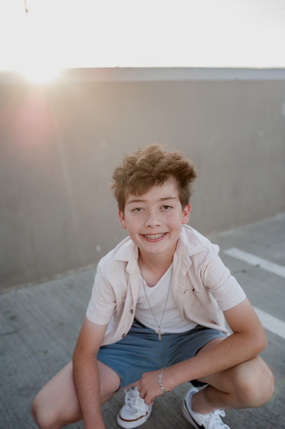 Smiling teenage boy with braces crouching on a rooftop parking lot at sunset, wearing a pink shirt and blue shorts.