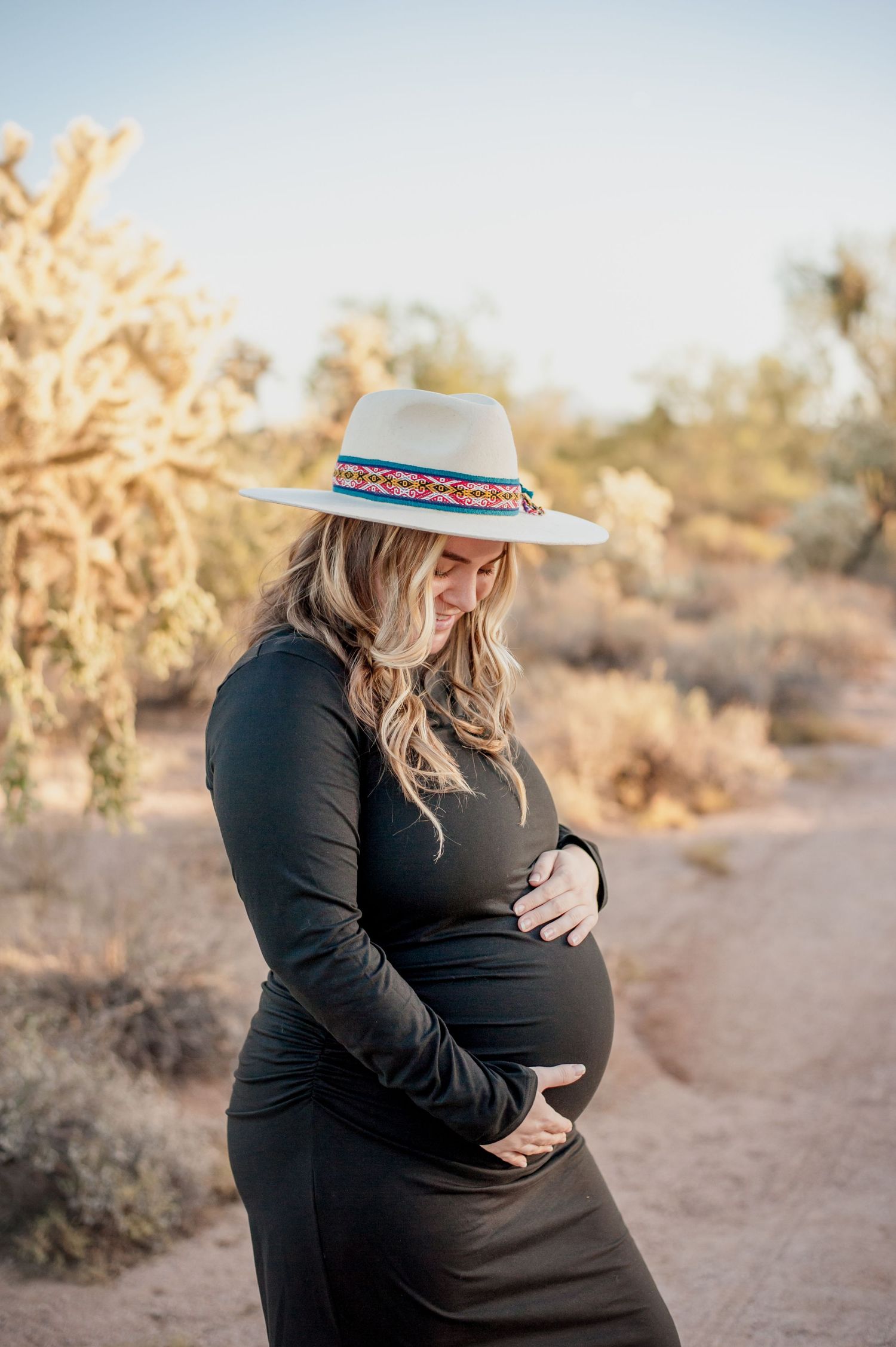 A pregnant woman in a black dress and sun hat poses in a desert landscape with cacti and golden sunlight.