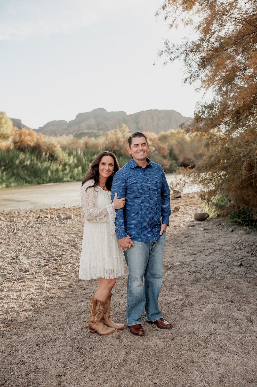 Couple smiling together near a desert river with mountain views, woman in white lace dress and cowboy boots, man in blue shirt.