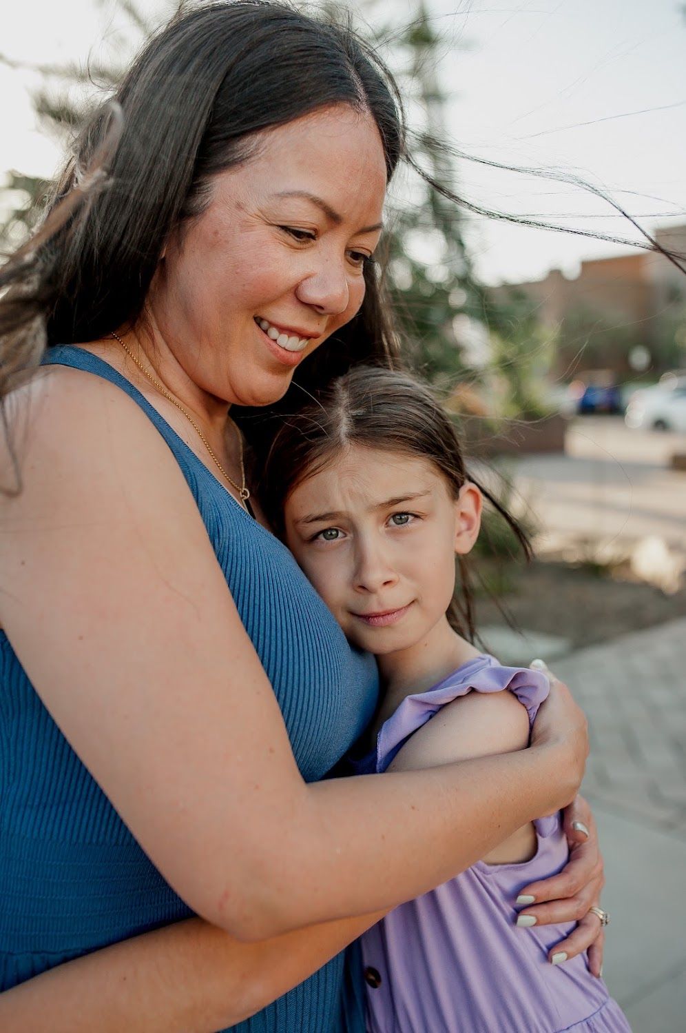 A woman in a blue dress hugs a young girl in lavender, both showing emotional expressions outdoors.