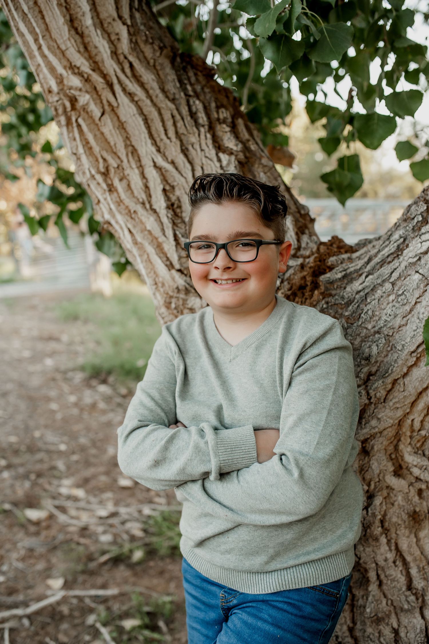 A young person in a gray sweater and blue jeans poses casually against a tree trunk in an outdoor setting.