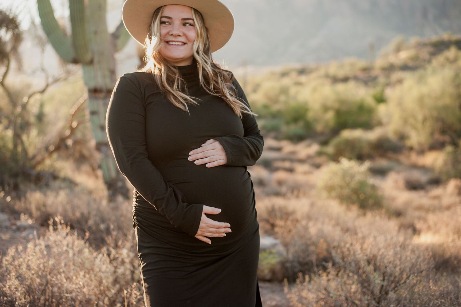 Expectant mother in dark dress and wide-brimmed hat poses in desert landscape during golden hour.