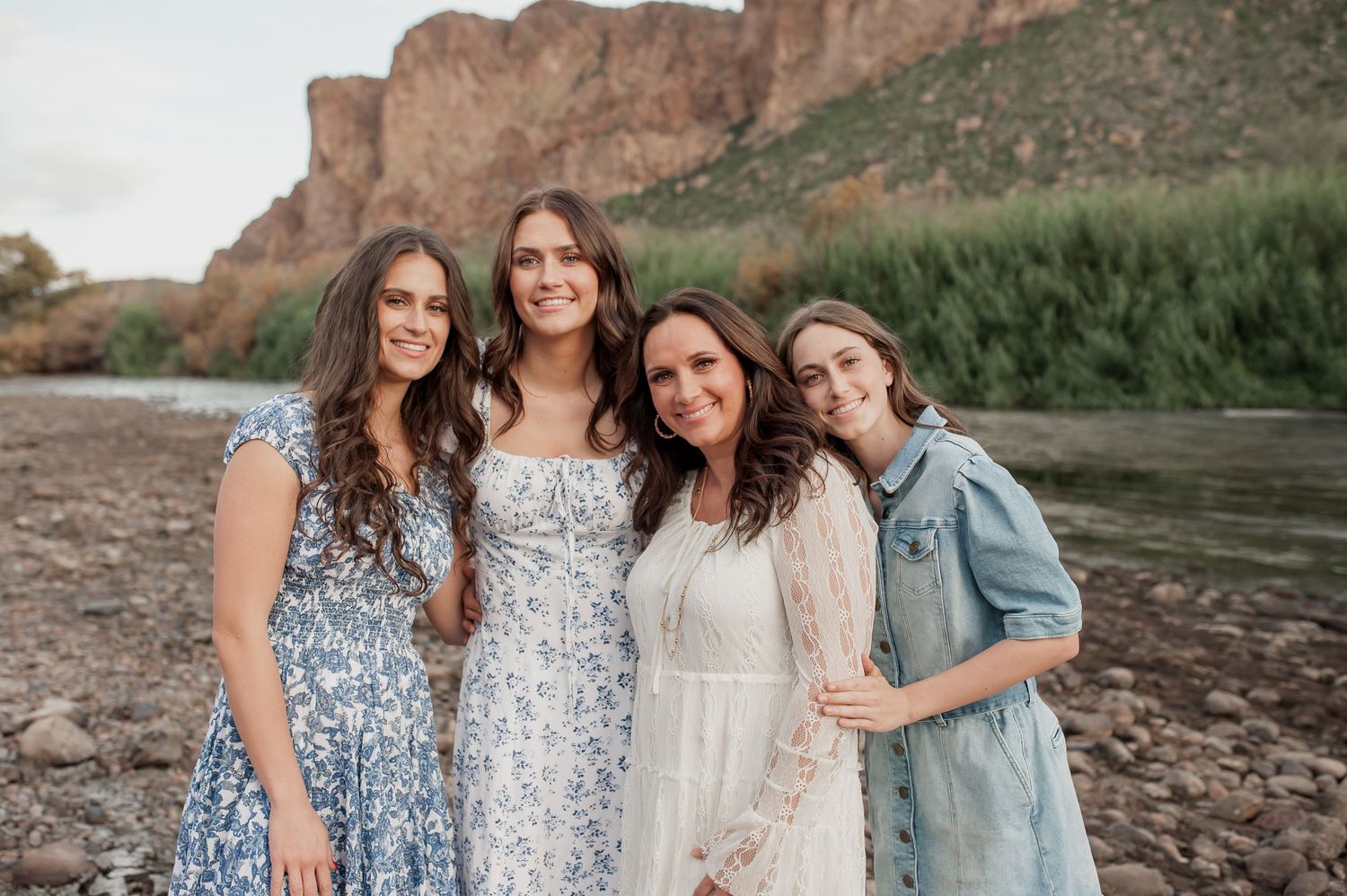 Group wearing coordinating blue and white dresses poses together on rocky beach near river at sunset.