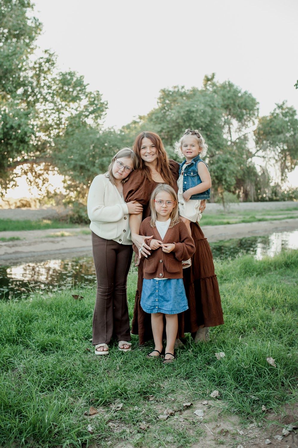 A woman and three young girls pose together outdoors near a pond surrounded by green trees and grass.