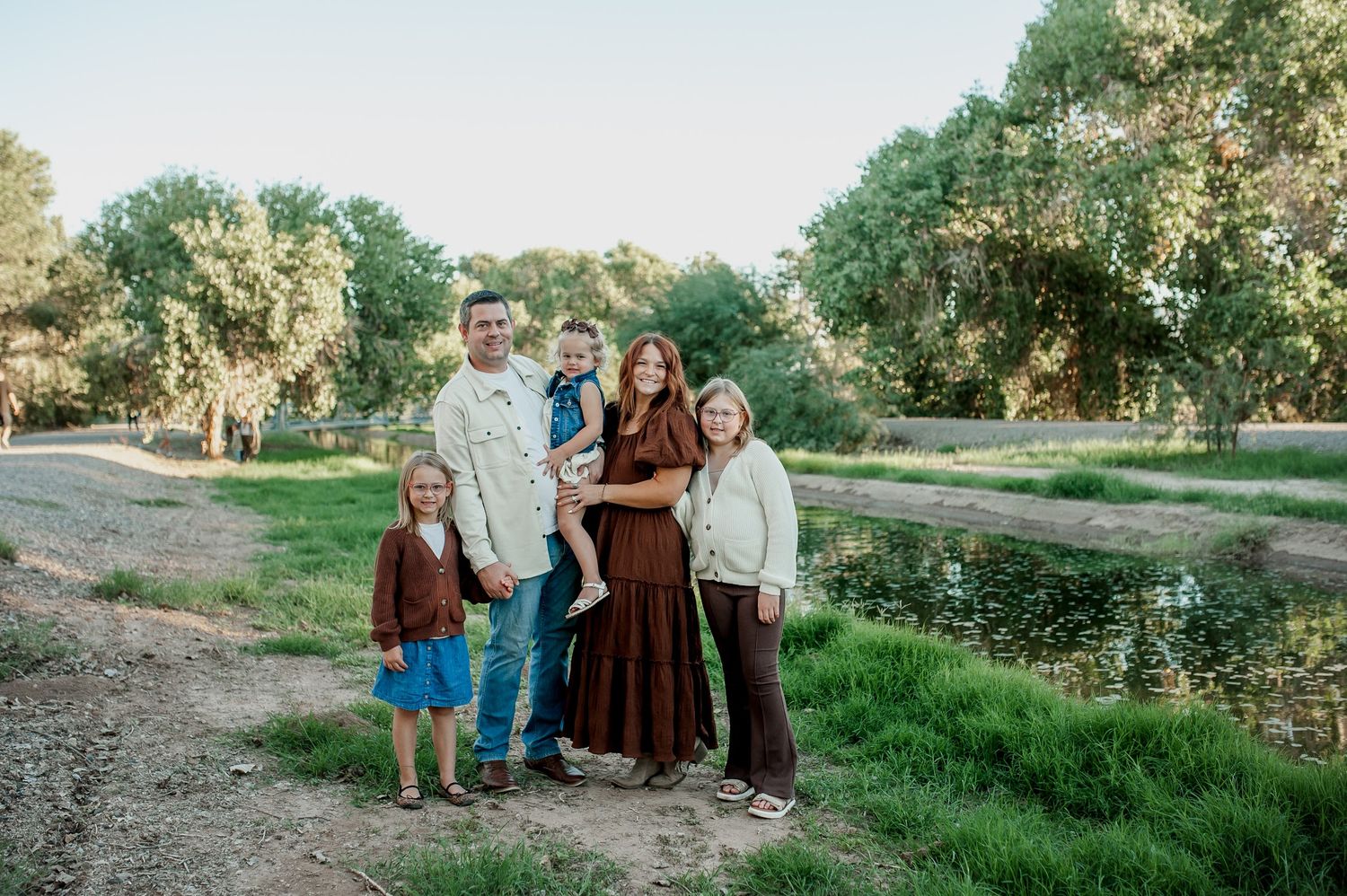 Family of five posing outdoors near a pond surrounded by lush green trees in a park setting.