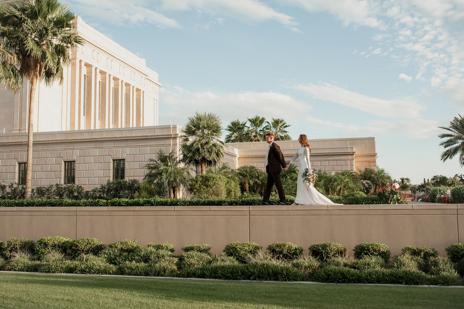 Bride and groom walking hand in hand outside a white temple surrounded by palm trees and lush greenery on their wedding day.