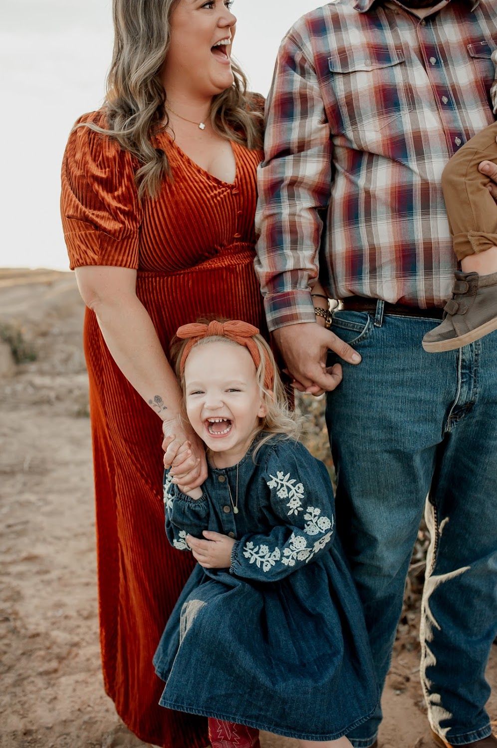 A joyful family photo with a laughing toddler girl in a denim dress holding hands with her mom in a rust velvet dress.