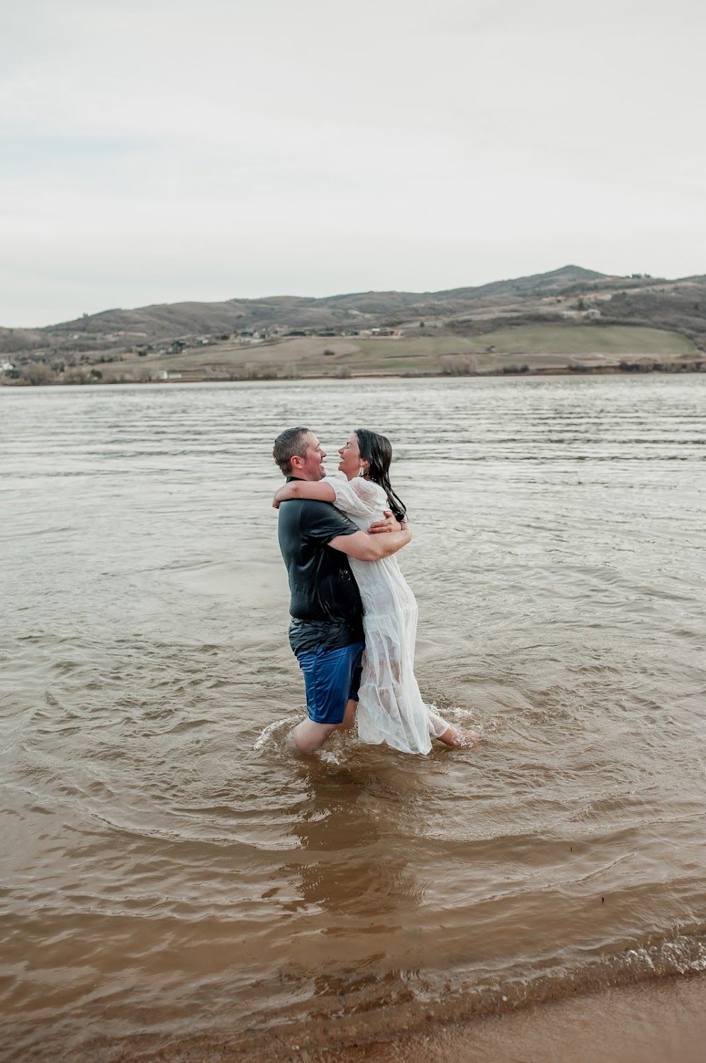 Couple embracing romantically while standing in shallow lake water, woman in white dress, hills in background.