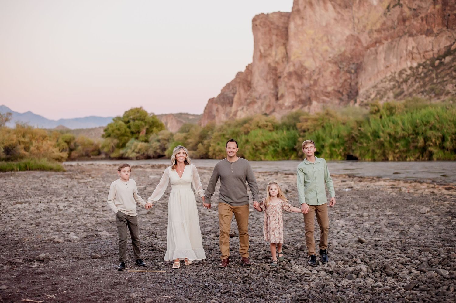 Family of five holding hands on rocky riverbank with desert canyon cliffs at sunset in Arizona.
