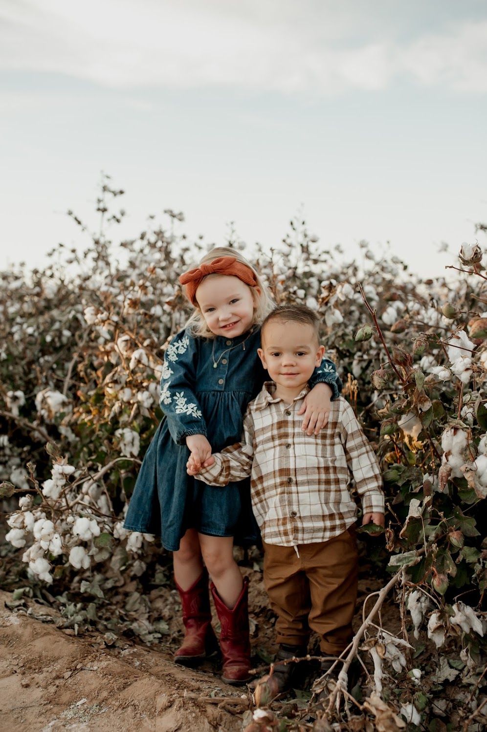 Two young children pose in a cotton field, girl in denim dress and red boots, boy in plaid shirt and tan pants.