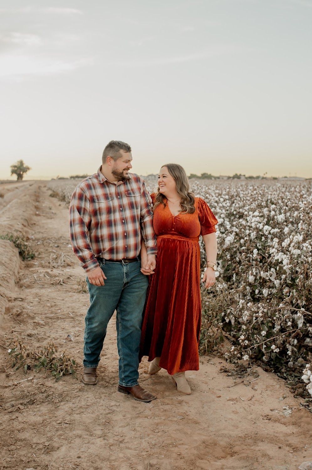 Couple holding hands smiling in a cotton field, woman in rust dress, man in plaid shirt and jeans, golden hour light.