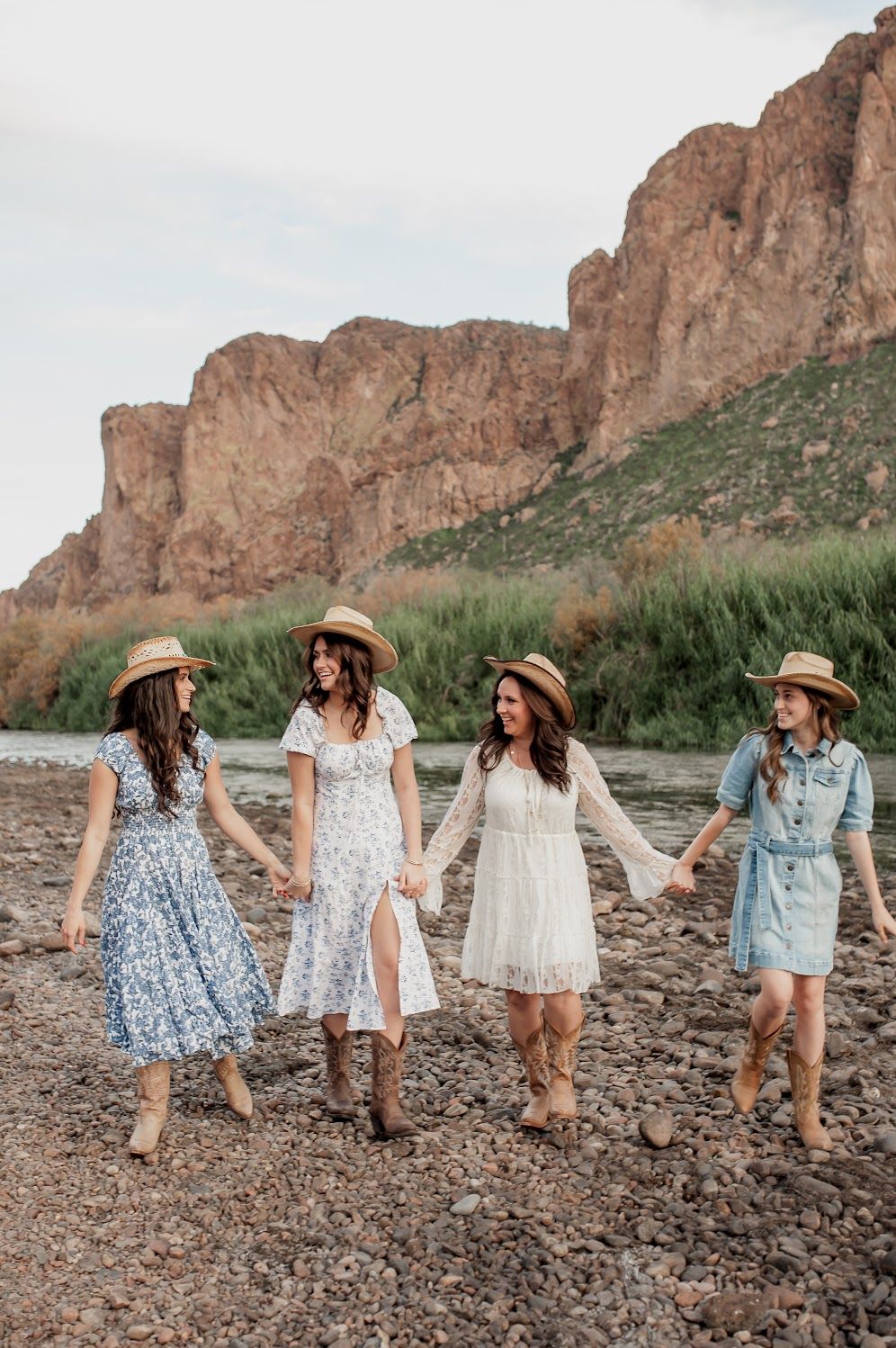 Four women in straw hats and dresses hold hands walking on rocky terrain with red cliffs and greenery behind them.