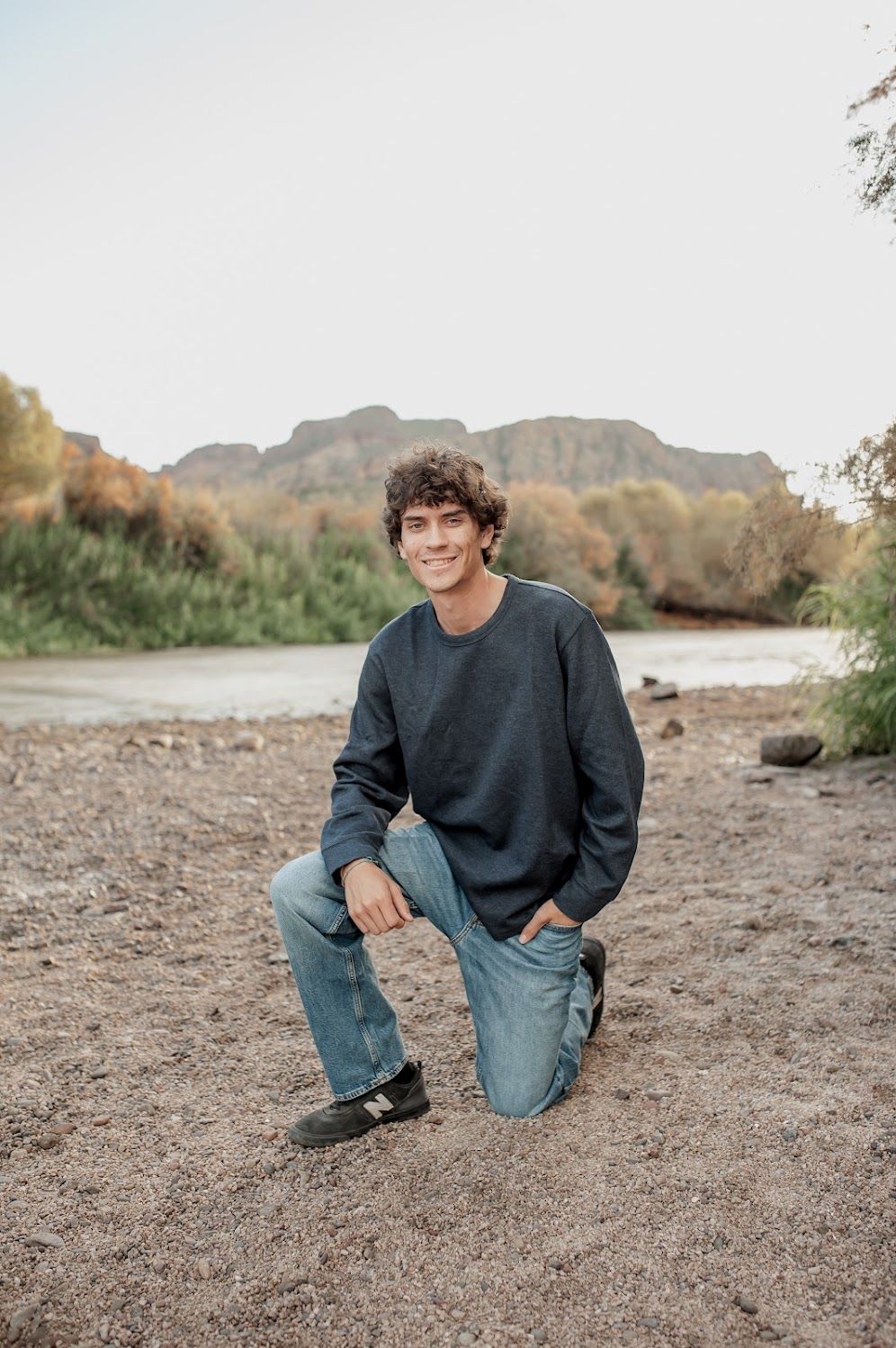 Young man with curly hair kneeling on sandy riverbank, wearing blue jeans and gray sweatshirt, with mountain backdrop.