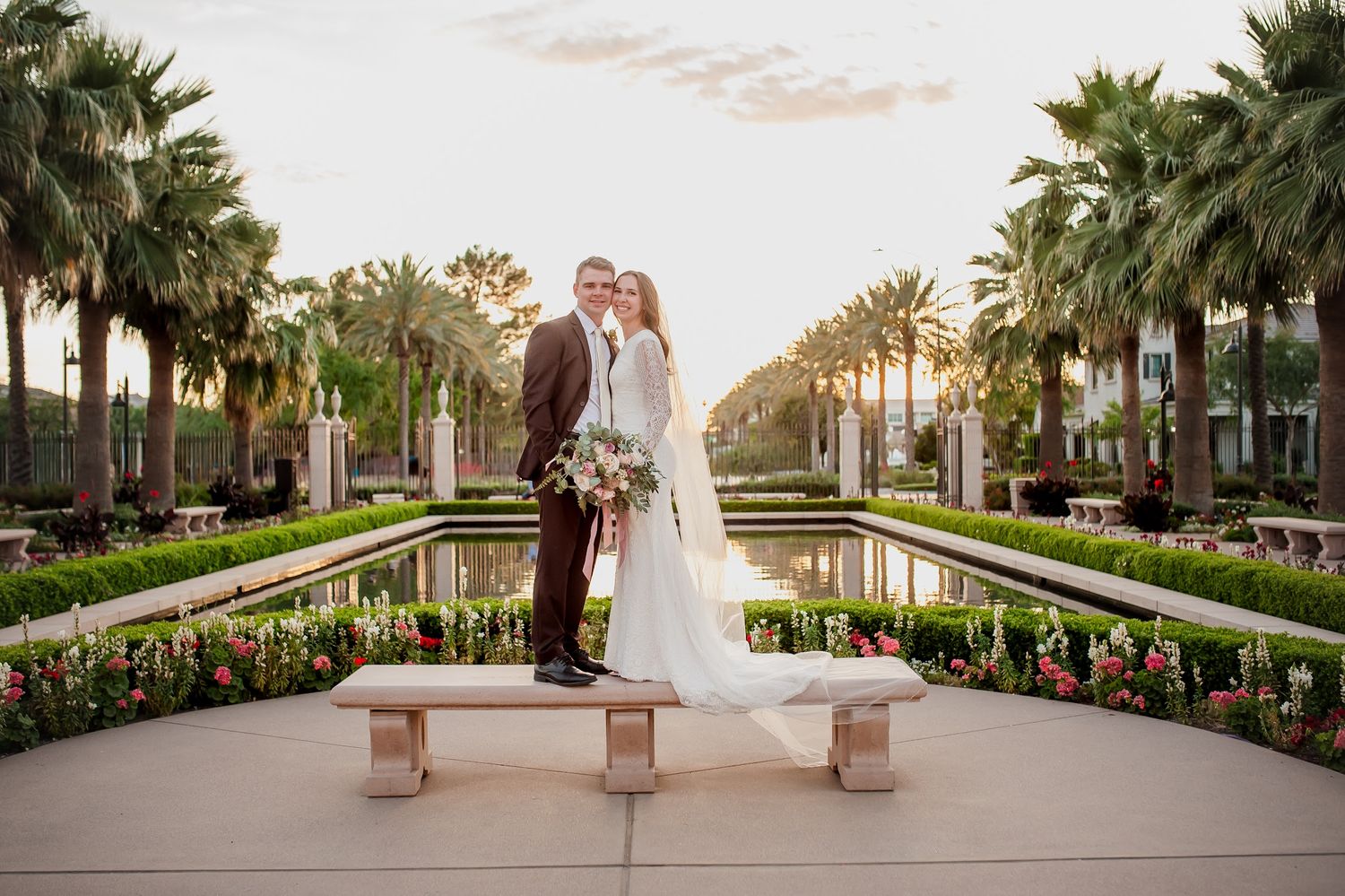 Bride and groom pose by a reflecting pool lined with palm trees and flowers at a elegant outdoor wedding venue.