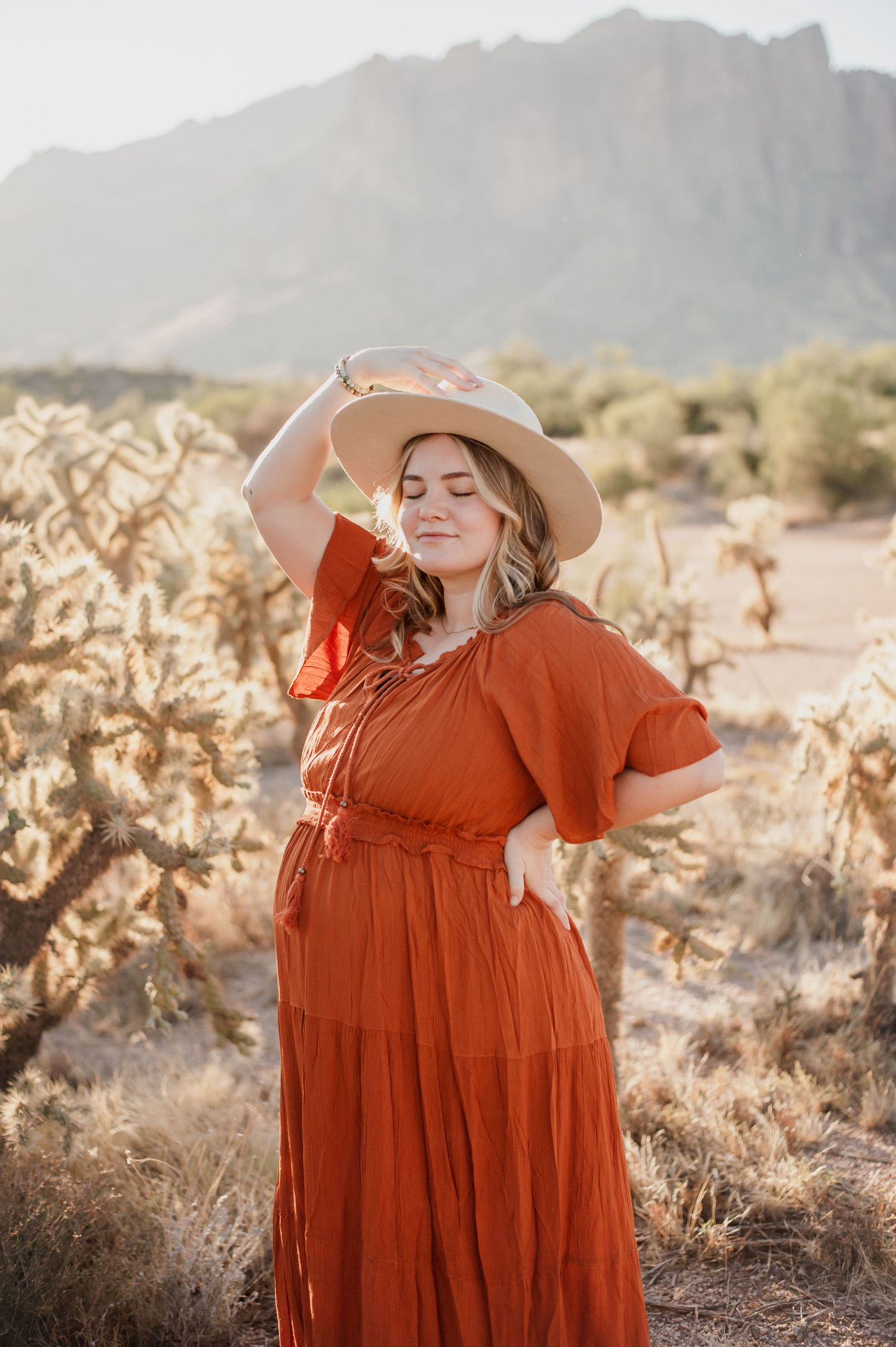 A woman in a rust-colored maxi dress and wide brim hat poses in a desert landscape with mountains in the background.
