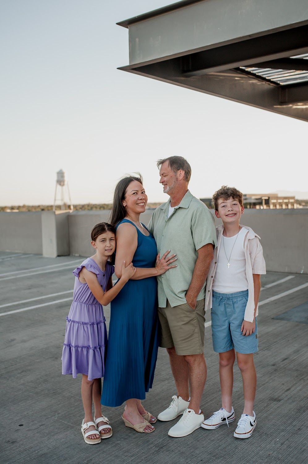 Happy family of four posing together on a rooftop parking deck at golden hour, smiling and embracing.