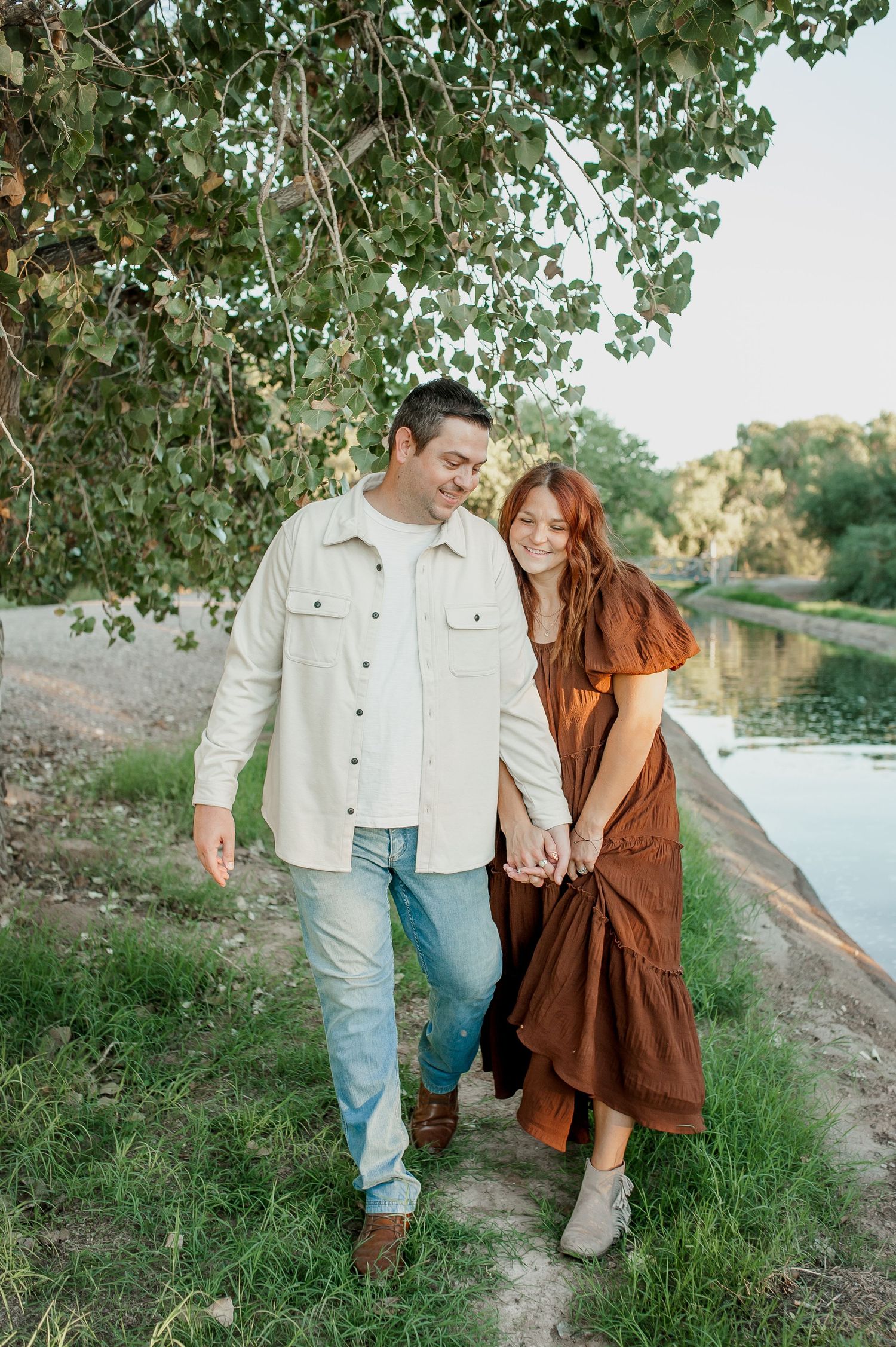 A couple holds hands and walks along a scenic riverside path beneath lush green trees during golden hour.