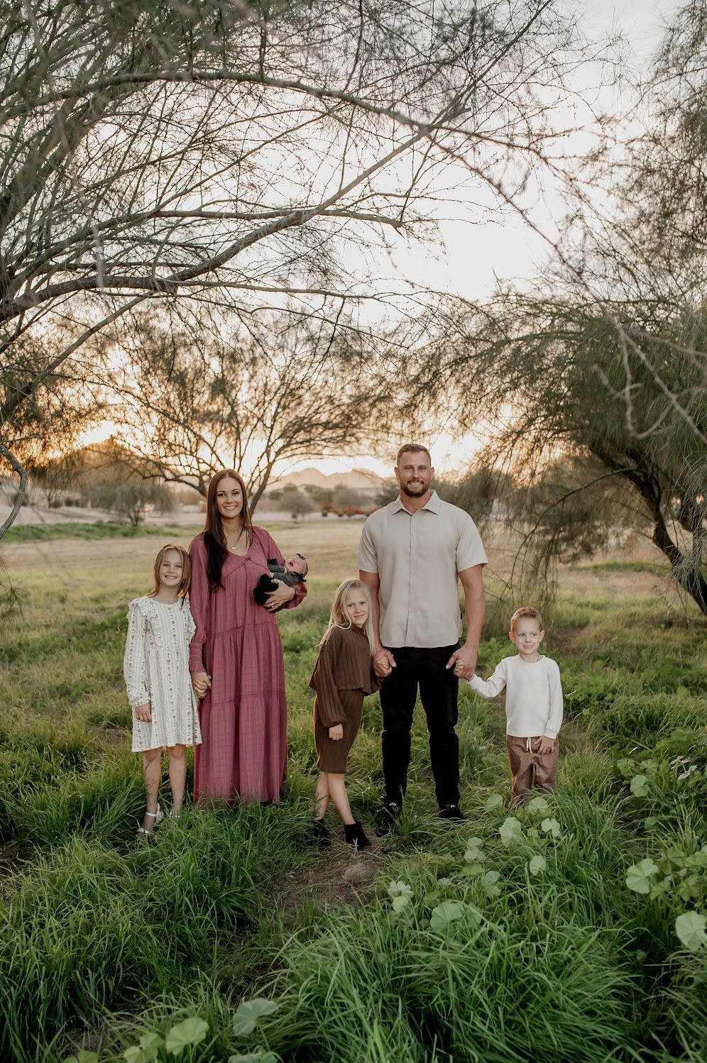 Family of five posing outdoors at golden hour among lush green grass and trees in a scenic park setting.