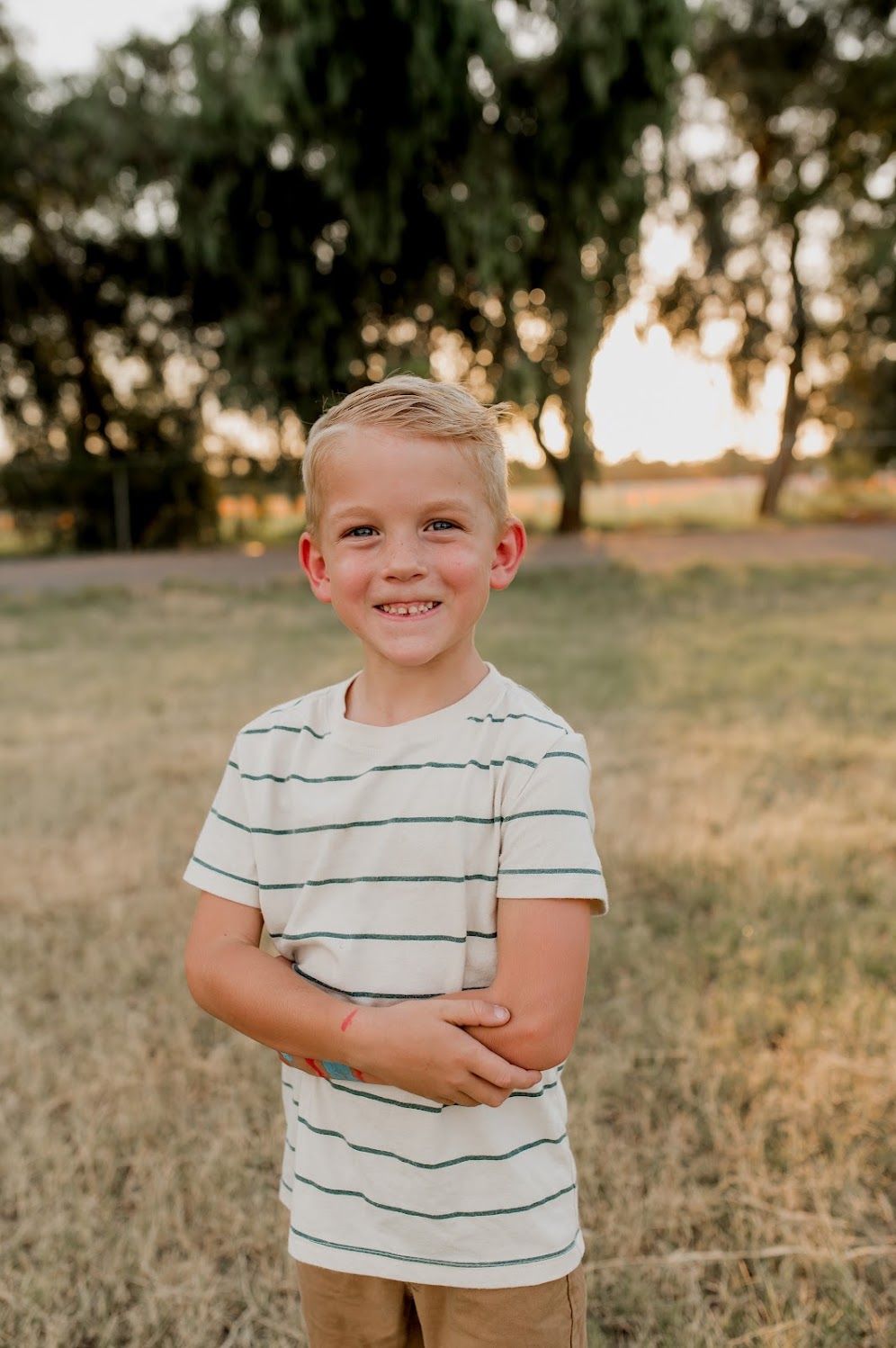 Smiling young boy in striped t-shirt standing in golden field with trees at sunset.