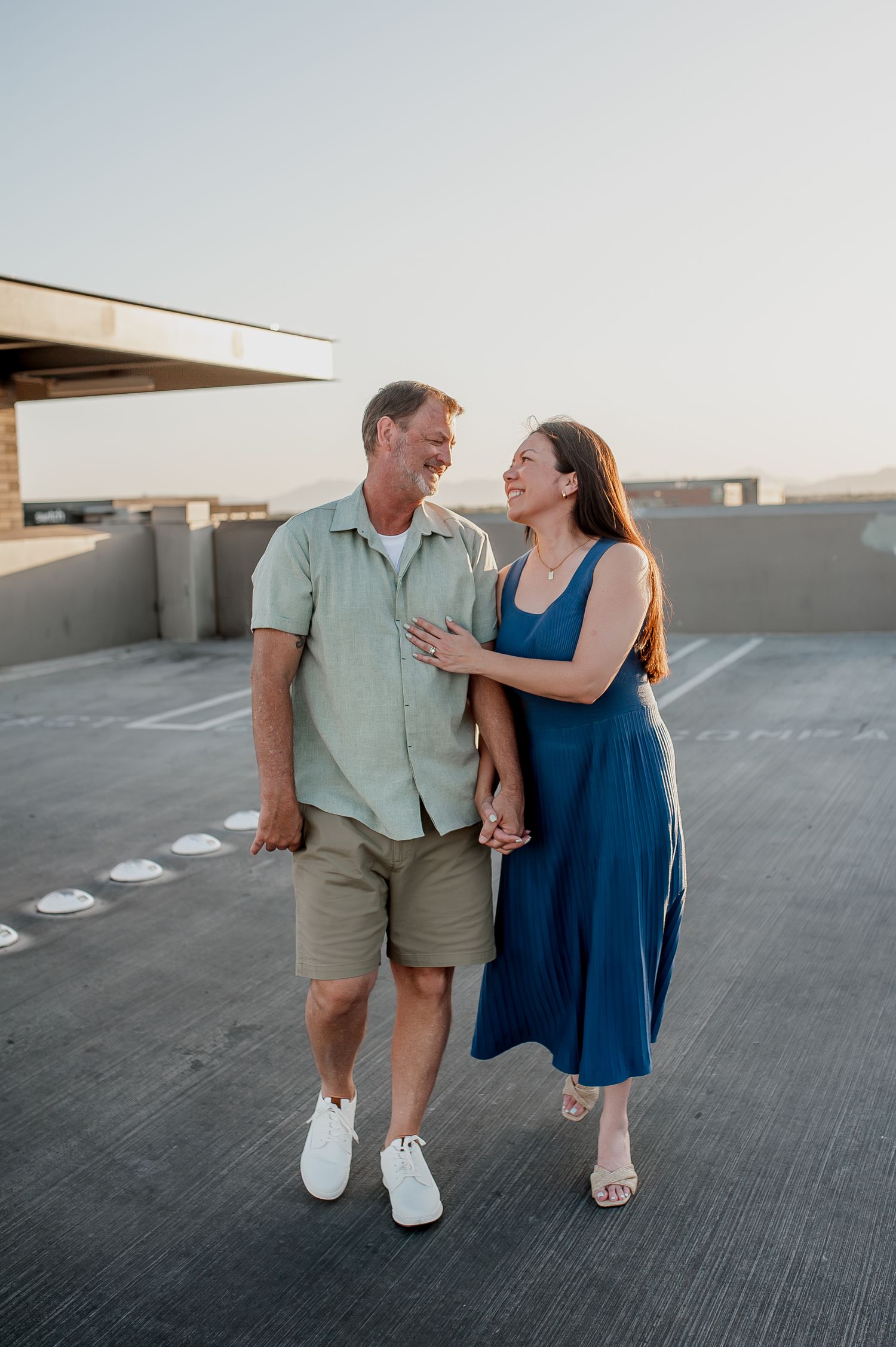 A smiling couple walks hand-in-hand on a rooftop parking lot at sunset, dressed in casual summer attire.