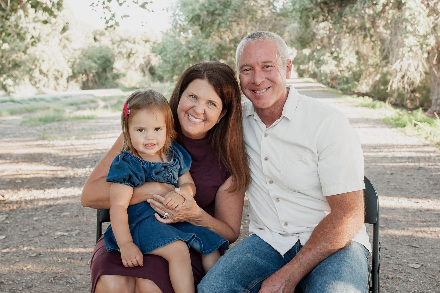 A family sits together outdoors on a bench along a tree-lined path on a sunny day.