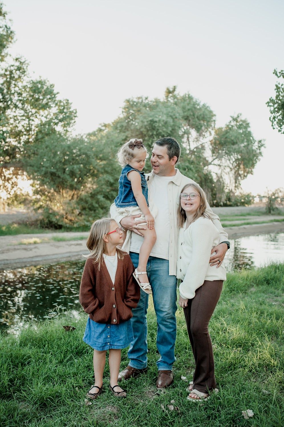 Family of four posing outdoors near a pond, with dad holding toddler, two girls in fall outfits, surrounded by green trees.
