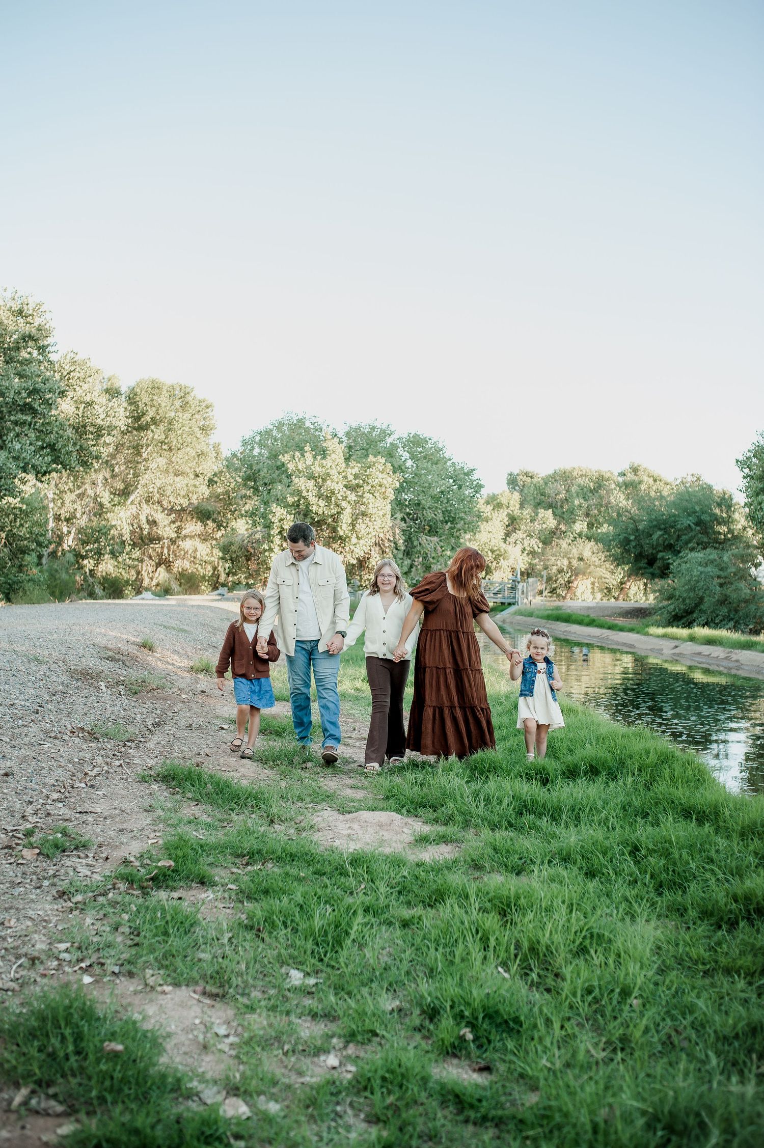 Family of five walking along a grassy riverbank path surrounded by lush green trees during golden hour.
