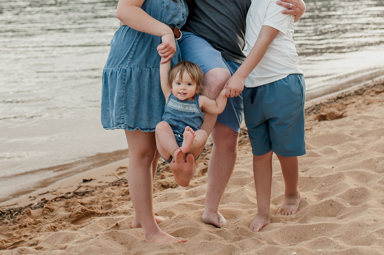 Family of three enjoying a sunny beach day, with parents playfully lifting their baby on golden sand near the water.
