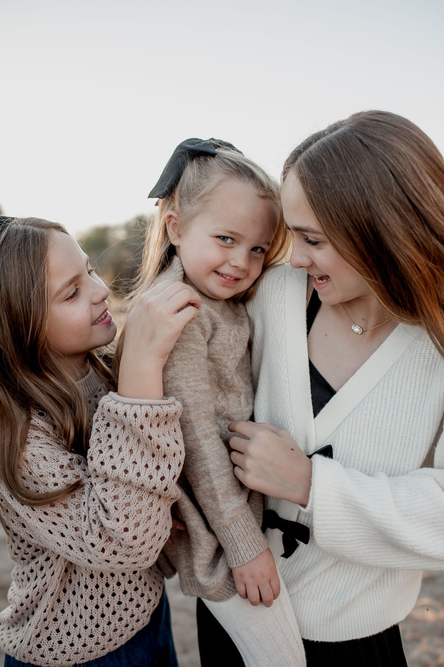 Two older sisters lovingly embrace their smiling toddler sister with a bow outdoors.