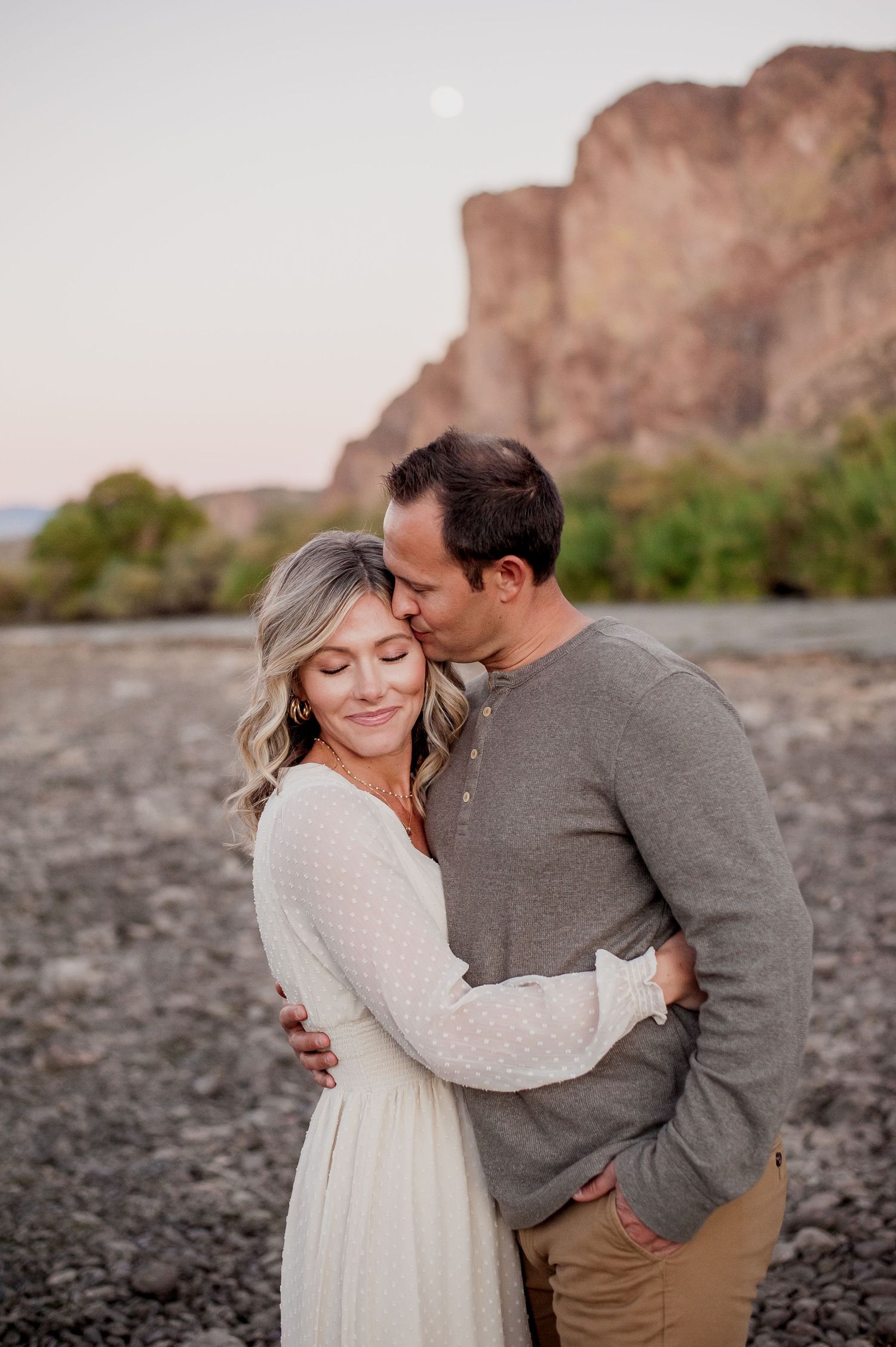 Couple embracing at sunset near rocky canyon, man kissing woman's temple as she smiles serenely.