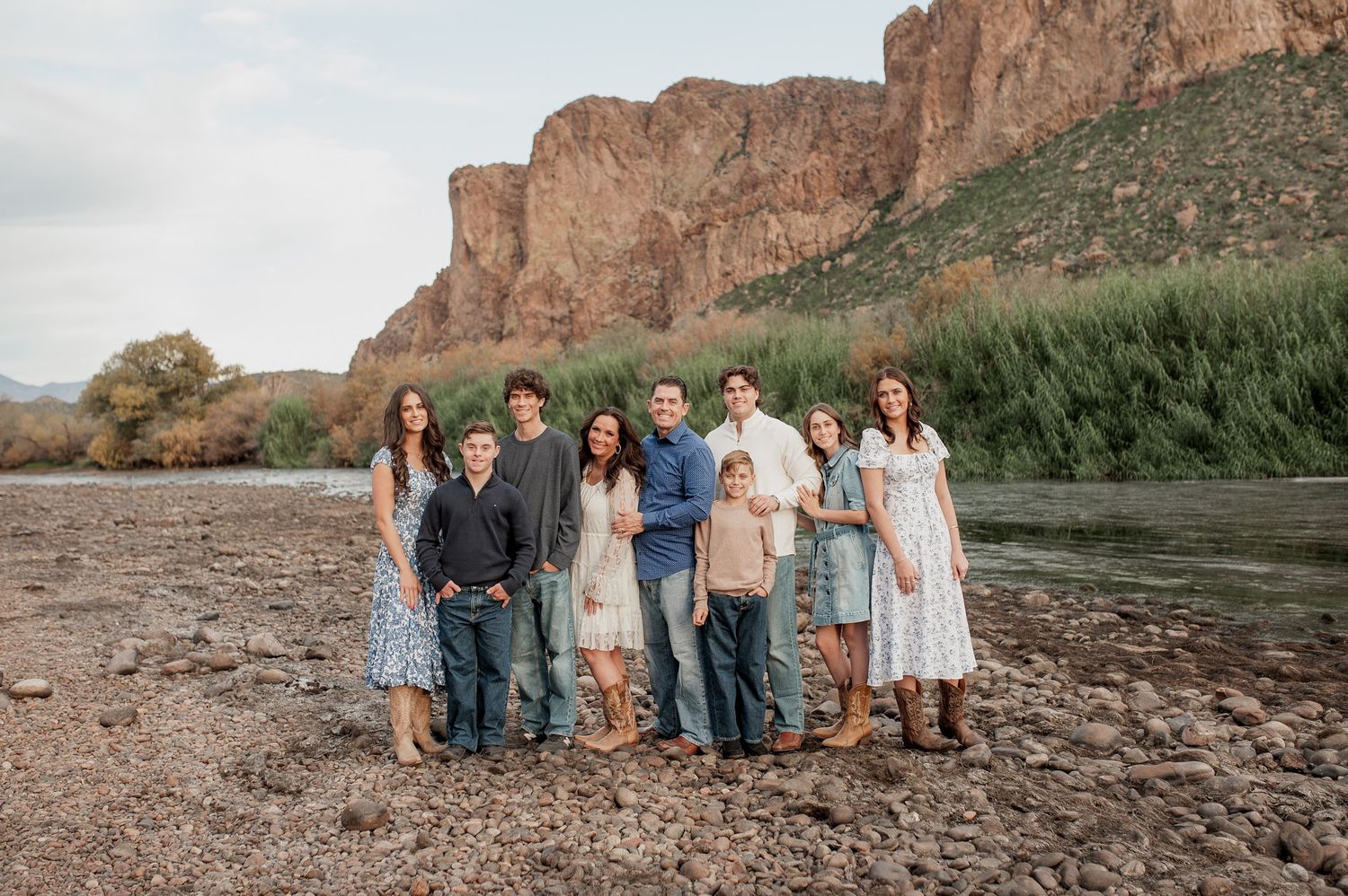 Large family posing together on rocky riverbank with red canyon cliffs and green reeds in background.