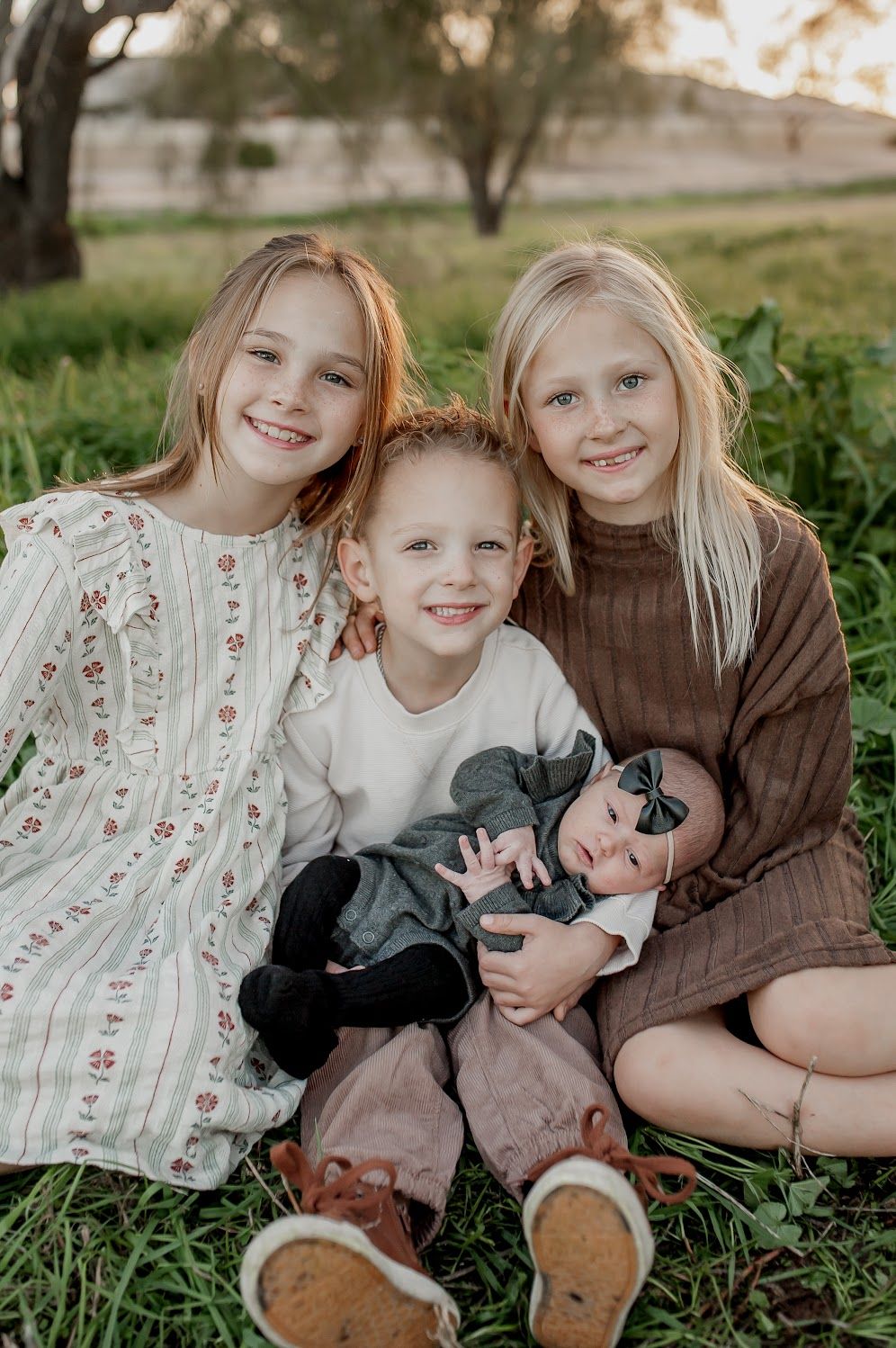 Three smiling children sit together on green grass at sunset, holding a sleeping newborn baby in a bow headband in their laps.