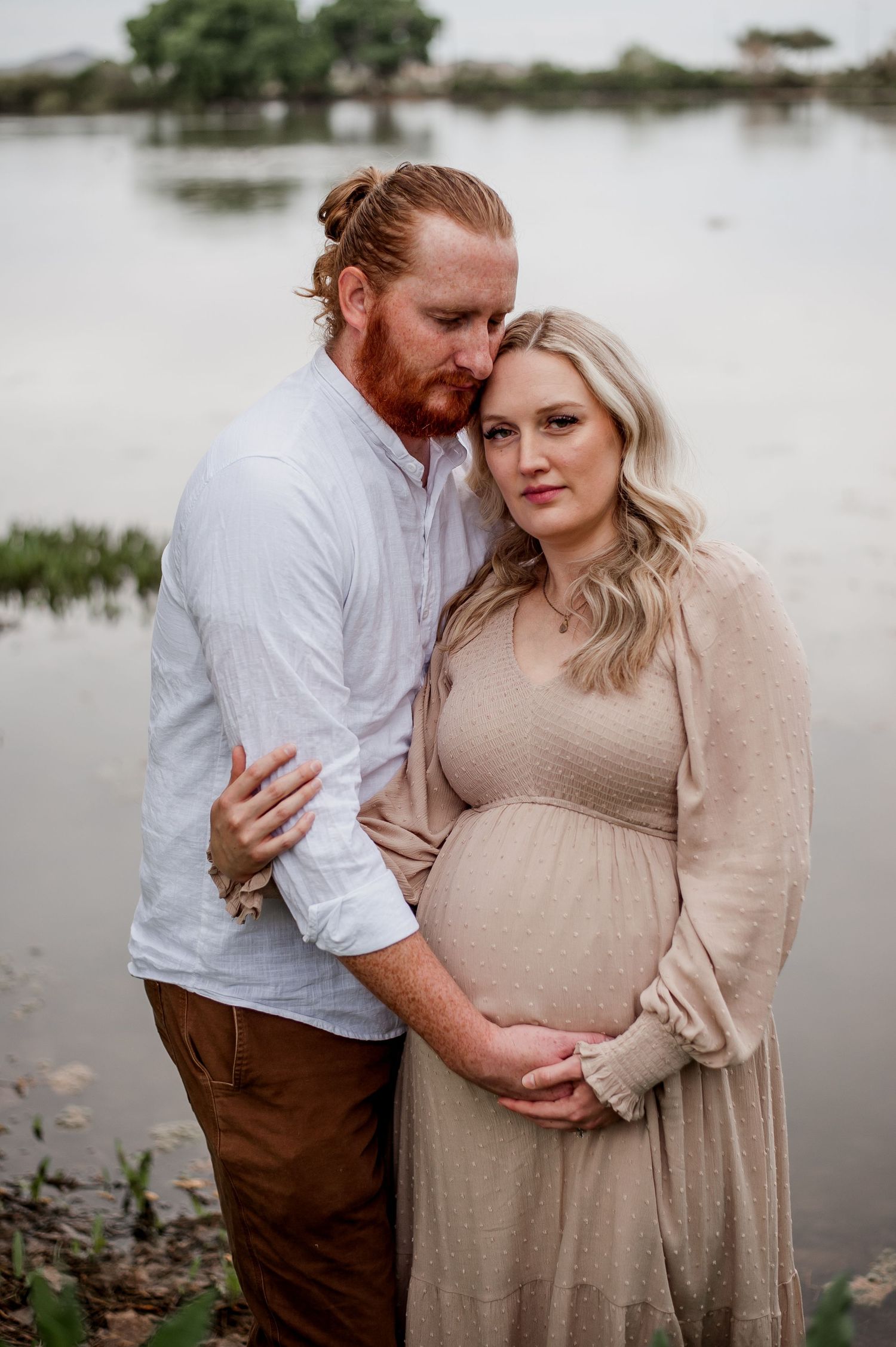 Romantic lakeside maternity portraits featuring couple in white and brown attire against water backdrop.