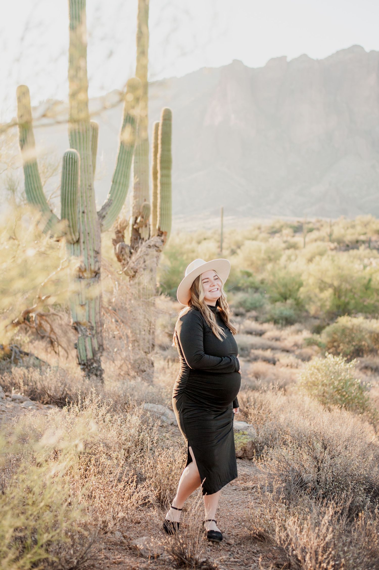 A figure in a black dress poses among saguaro cacti and desert brush during golden hour in the Arizona desert.