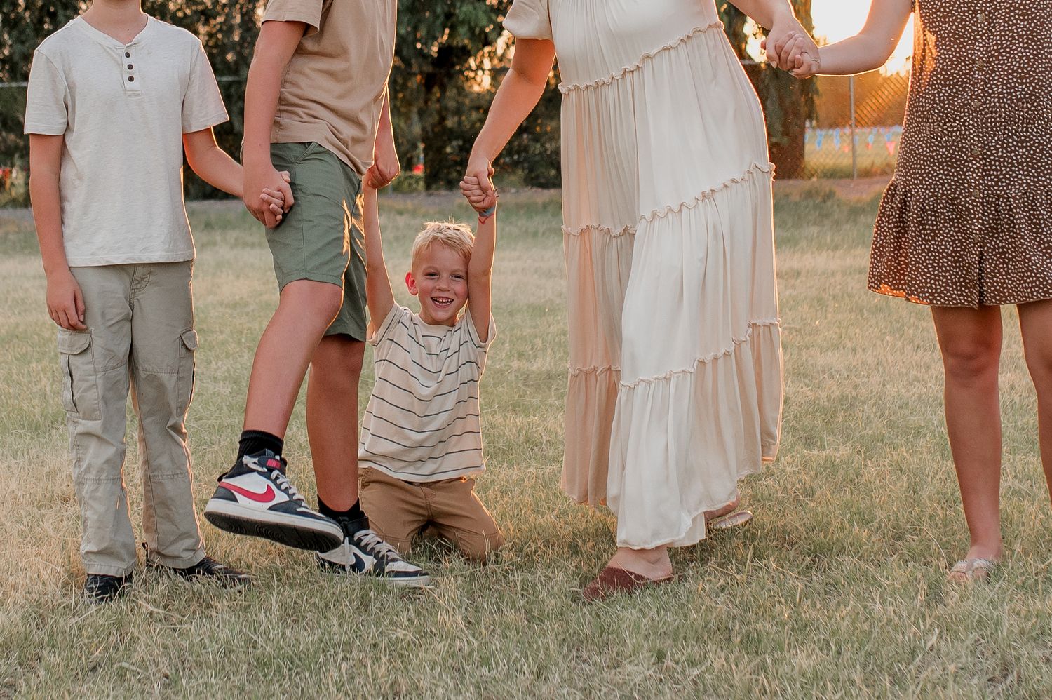 Family of four holding hands outdoors at golden hour, young child swinging between parents on green grass.