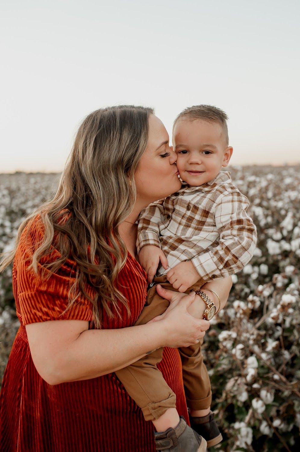 A mother kisses her smiling toddler boy while holding him in a cotton field during golden hour.