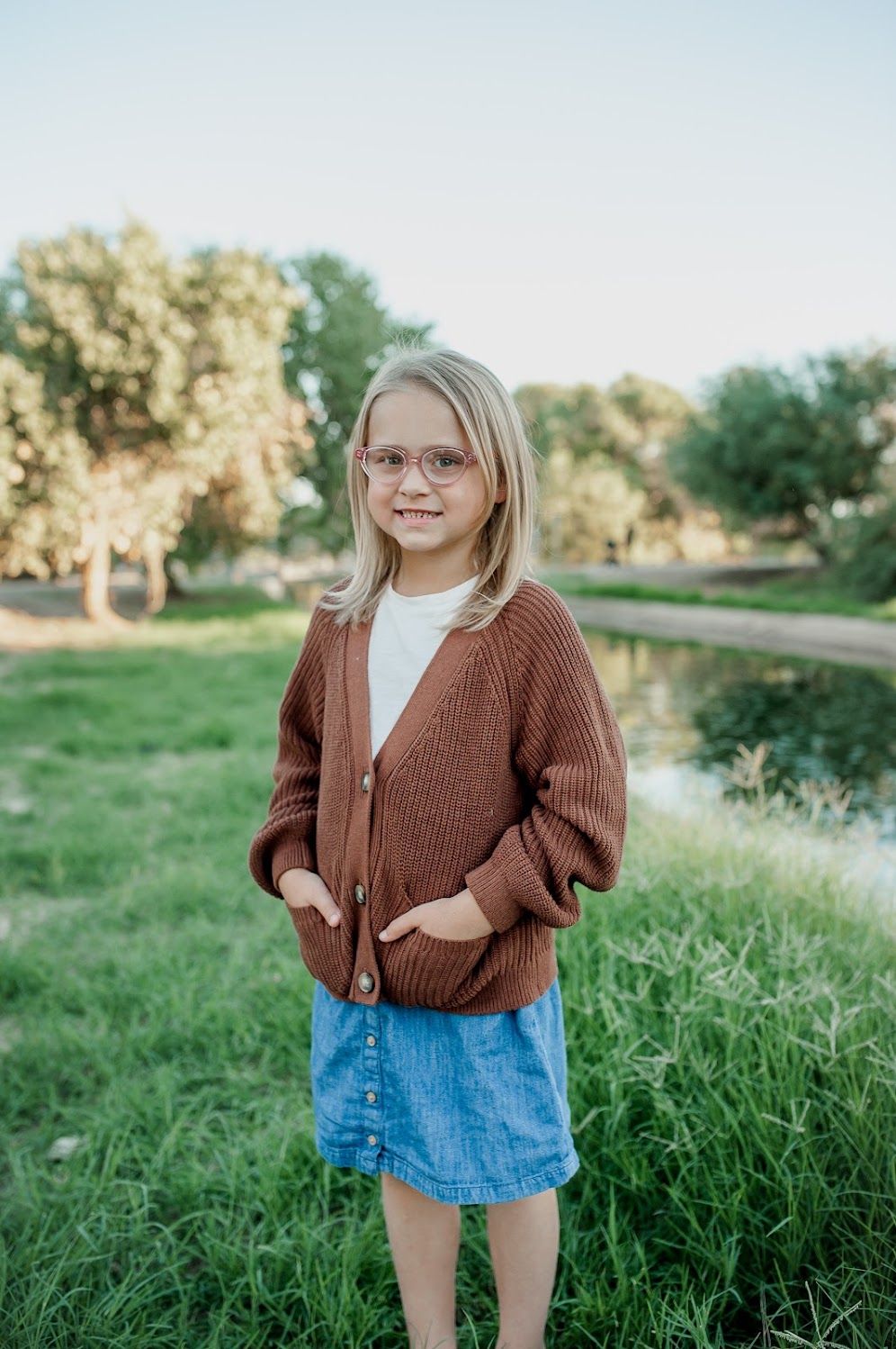 Young blonde girl in brown cardigan and denim skirt smiling outdoors in a green park setting.