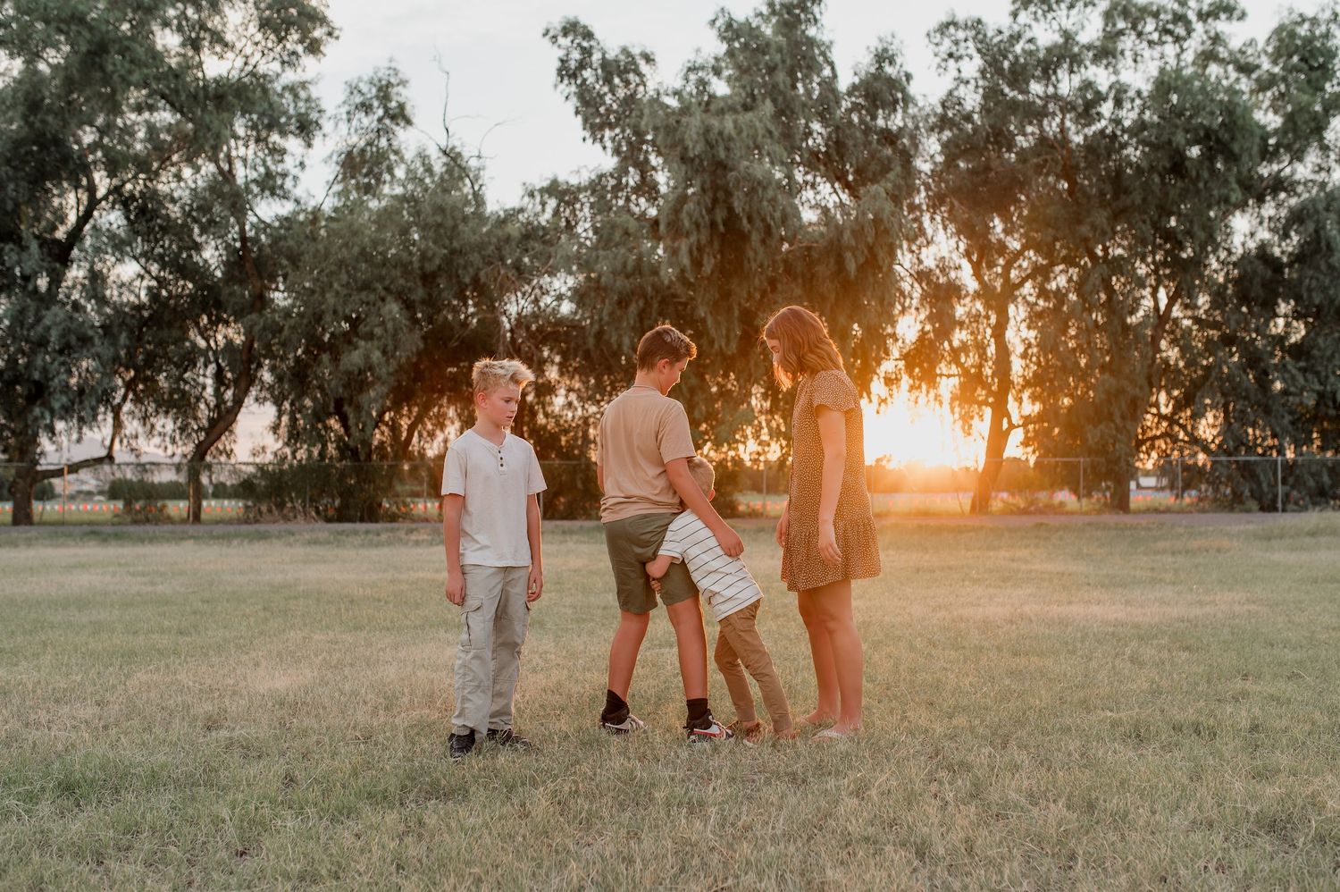 Four children stand together in a grassy field at golden sunset with large trees in the background.