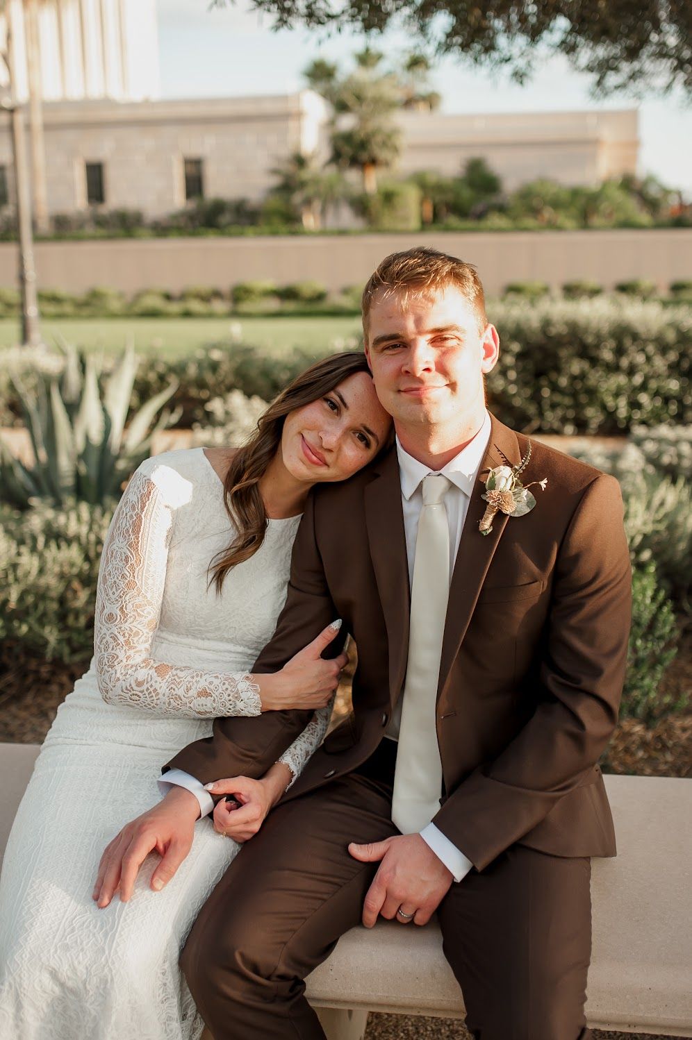 A bride in lace gown and groom in brown suit pose together outdoors at a wedding venue with gardens.