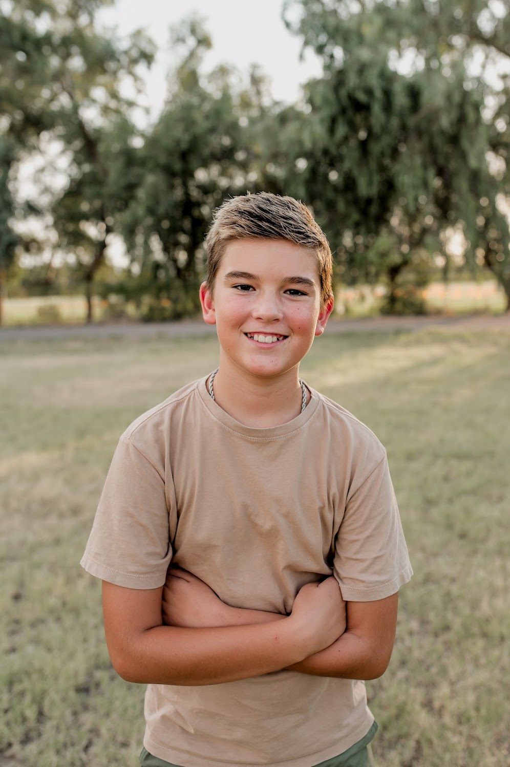 Smiling teenage boy with crossed arms wearing a tan t-shirt, standing outdoors in a green park setting.