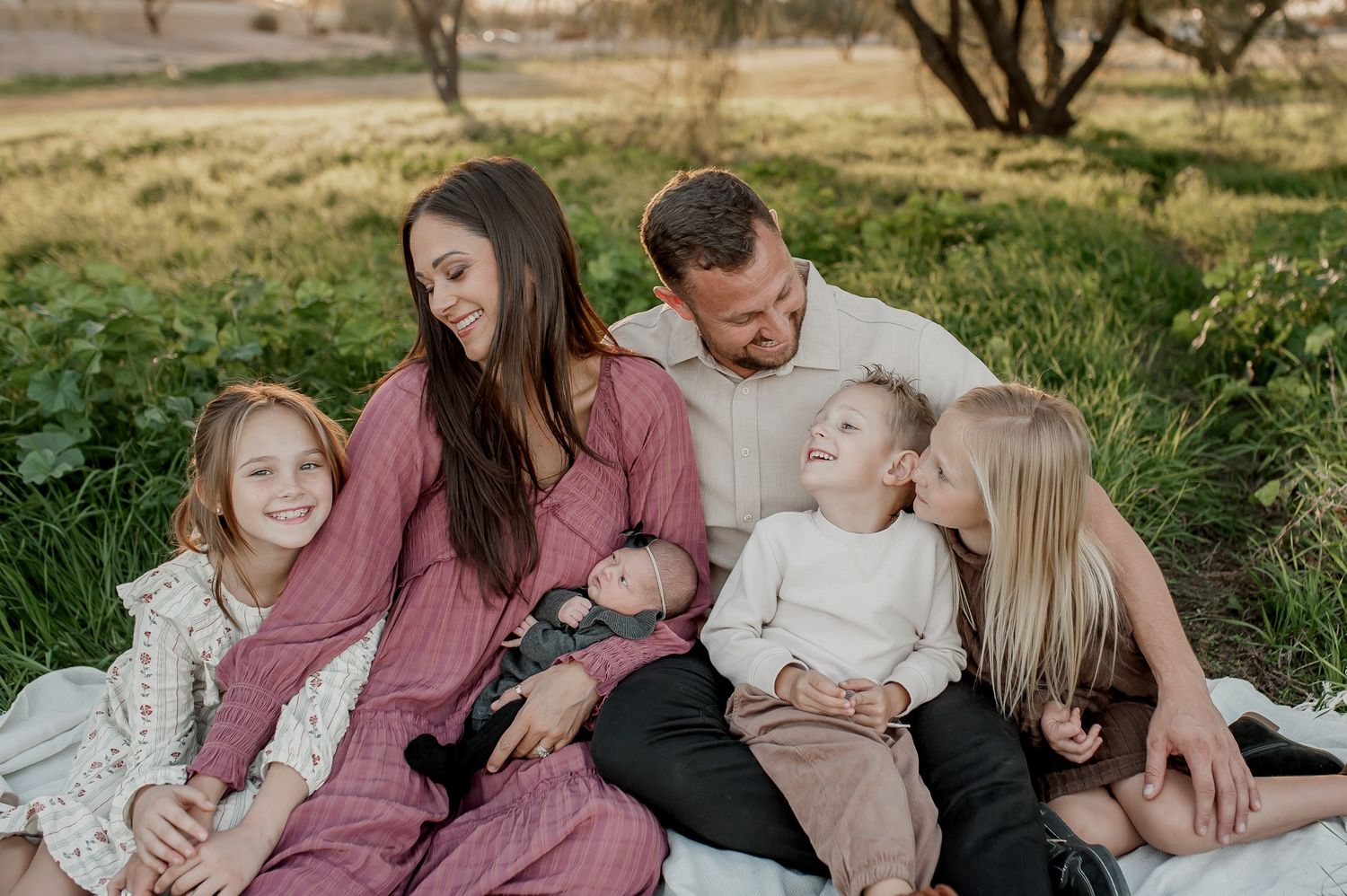 A happy family of six sits together outdoors on a blanket, parents smiling with four children including a newborn baby.