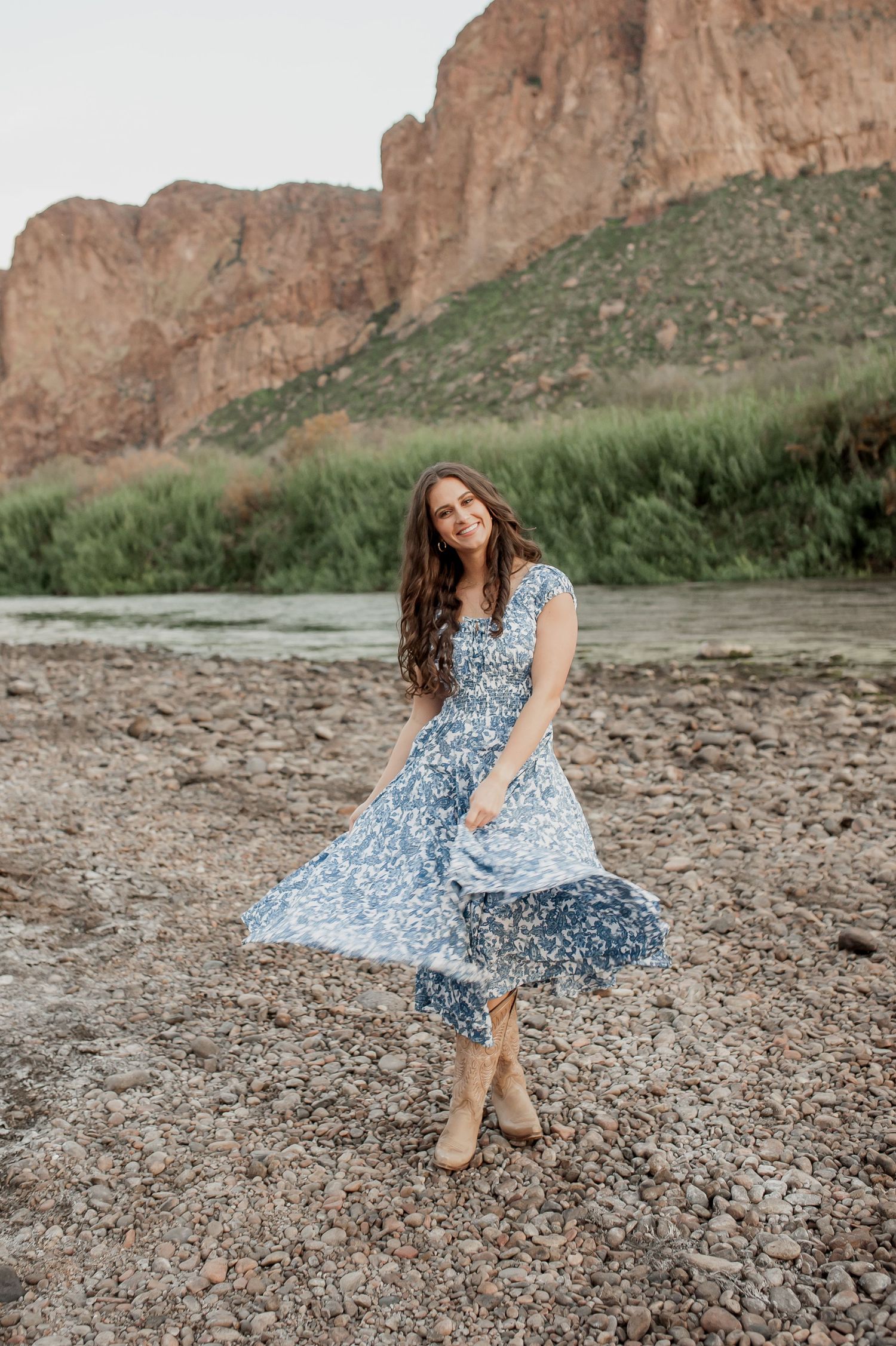 A flowing blue floral dress twirls gracefully against a backdrop of towering red rock cliffs and a winding river.