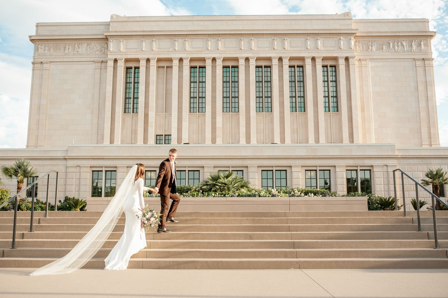 Bride and groom walk down steps of a grand white temple on their wedding day, with long veil trailing behind.